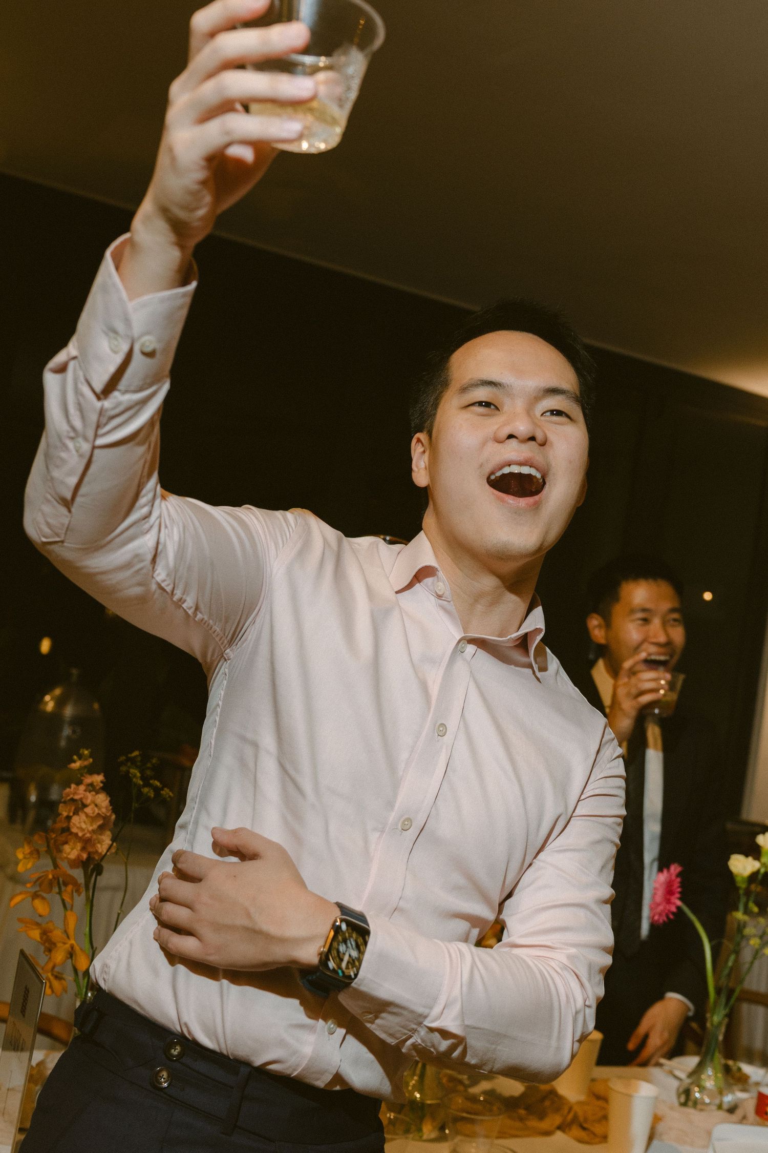 Wedding guest in white dress shirt celebrates enthusiastically with raised arms at reception party.