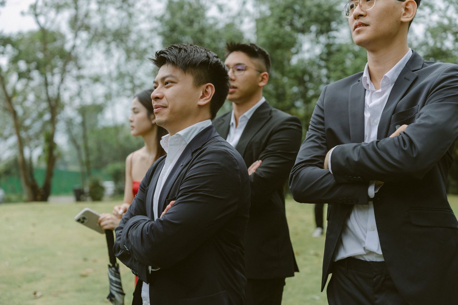 Groomsmen in black suits laugh and joke together during outdoor wedding photos in a natural setting.