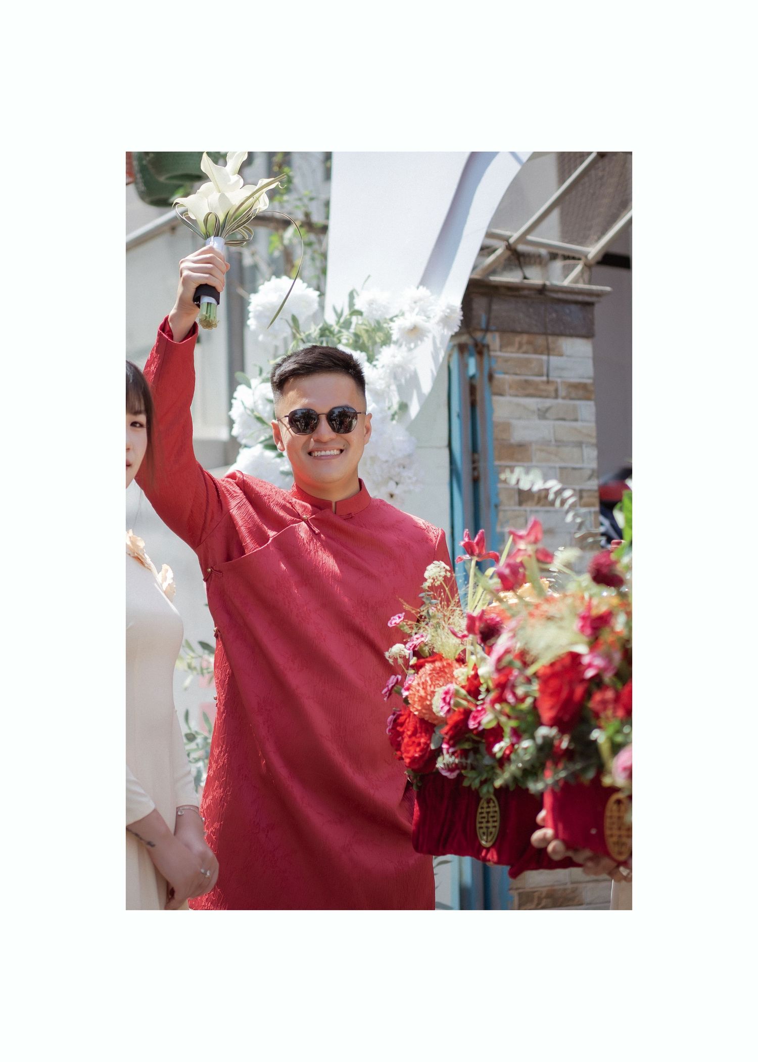 Person wearing red kurta with sunglasses smiles and poses with flowers in bright outdoor setting at decorative doorway.