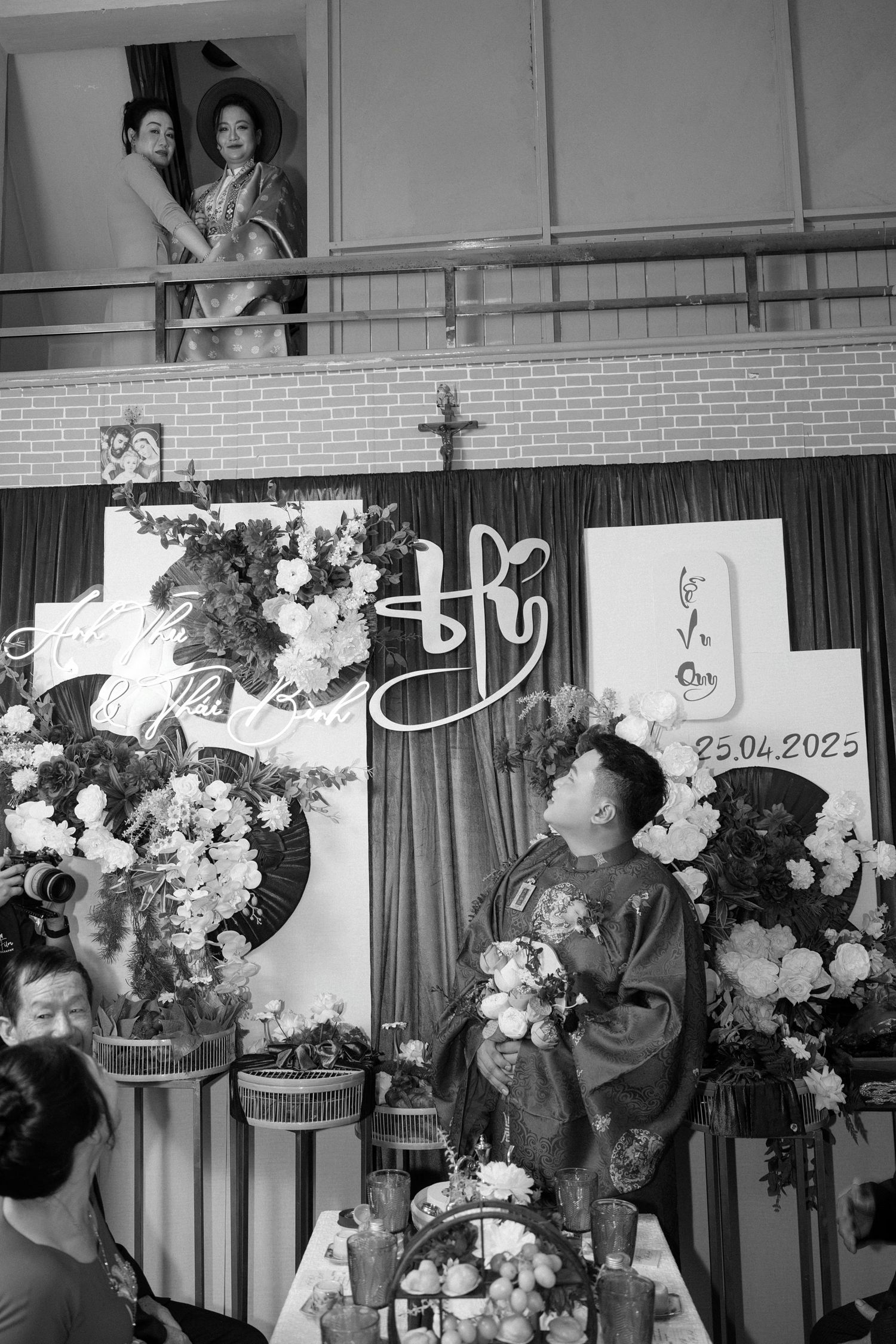 A black and white wedding photo showing guests gathered around floral arrangements and decorative displays at a venue.