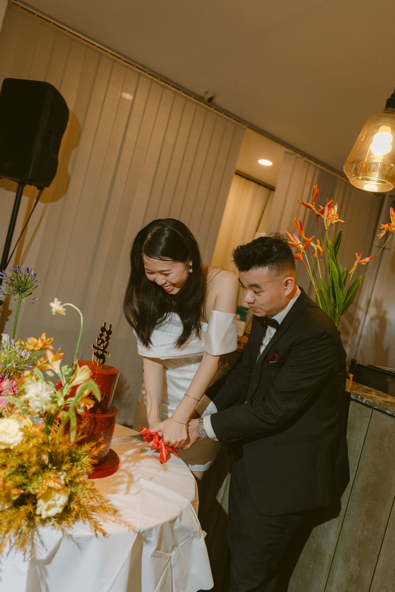 A couple in formal attire cuts their wedding cake together beside beautiful floral arrangements and decorative lighting.