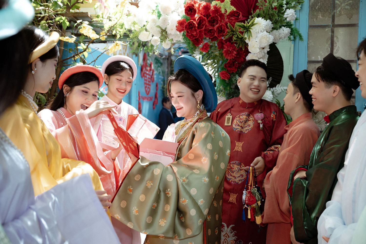 A group wearing traditional Asian hanbok clothing gather for a celebration with red flowers in the background.