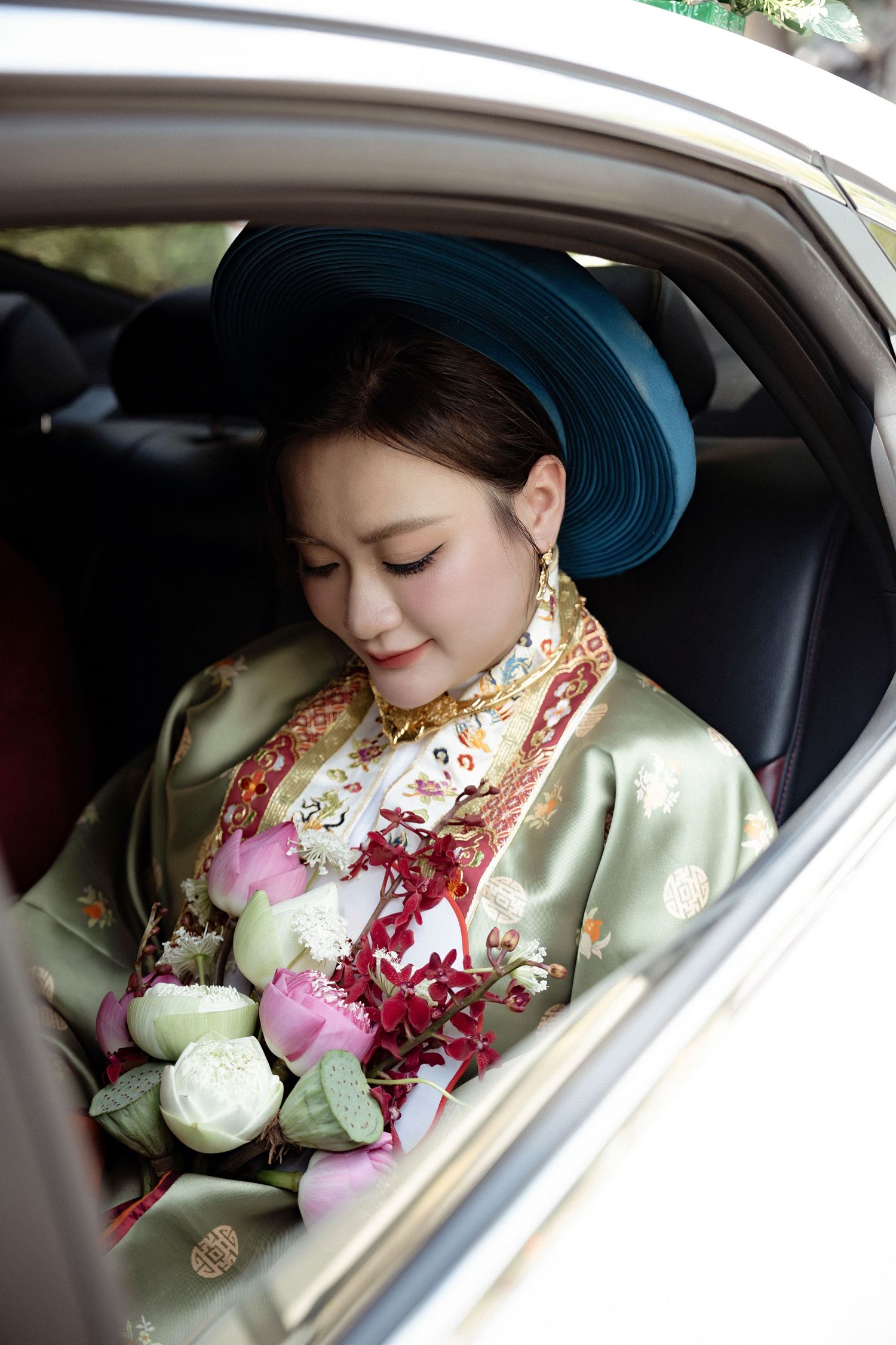 A traditional Vietnamese bride in an ornate ao dai and turquoise hat sits in a car with pink and white lotus flowers.