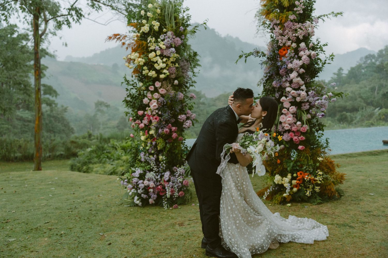 A romantic outdoor wedding ceremony under floral arches overlooking a misty mountain lake landscape.