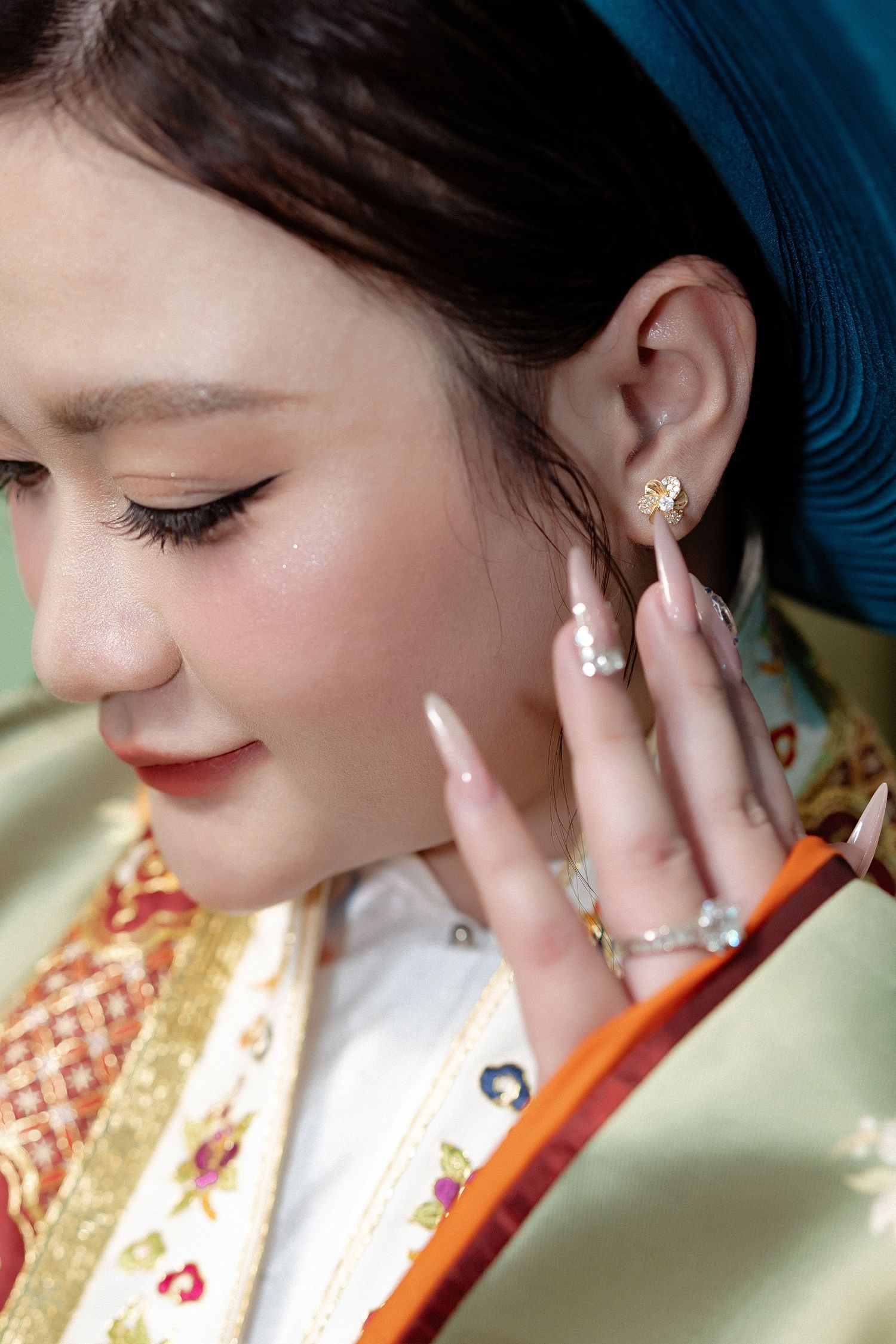 Close up detail of a hand with pink manicured nails touching delicate pearl earrings against traditional patterned fabric.