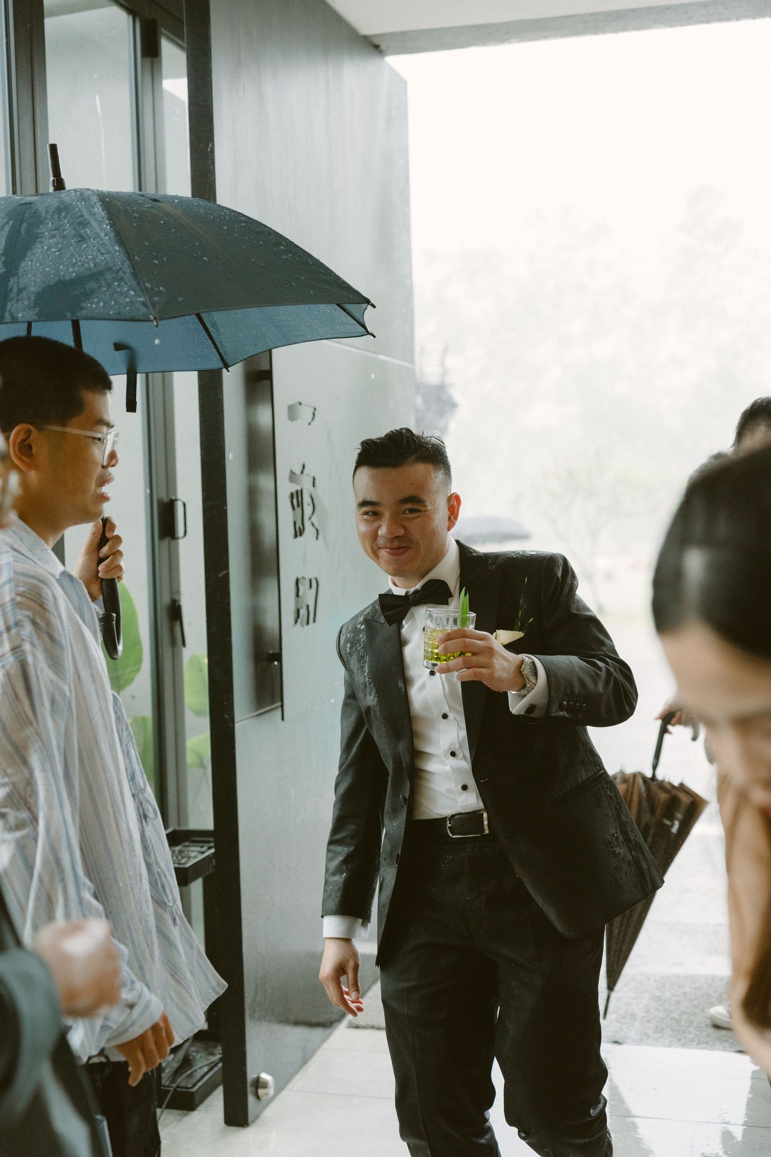 A wedding party stands under a blue umbrella in formalwear, with the guests sharing drinks during an outdoor reception.