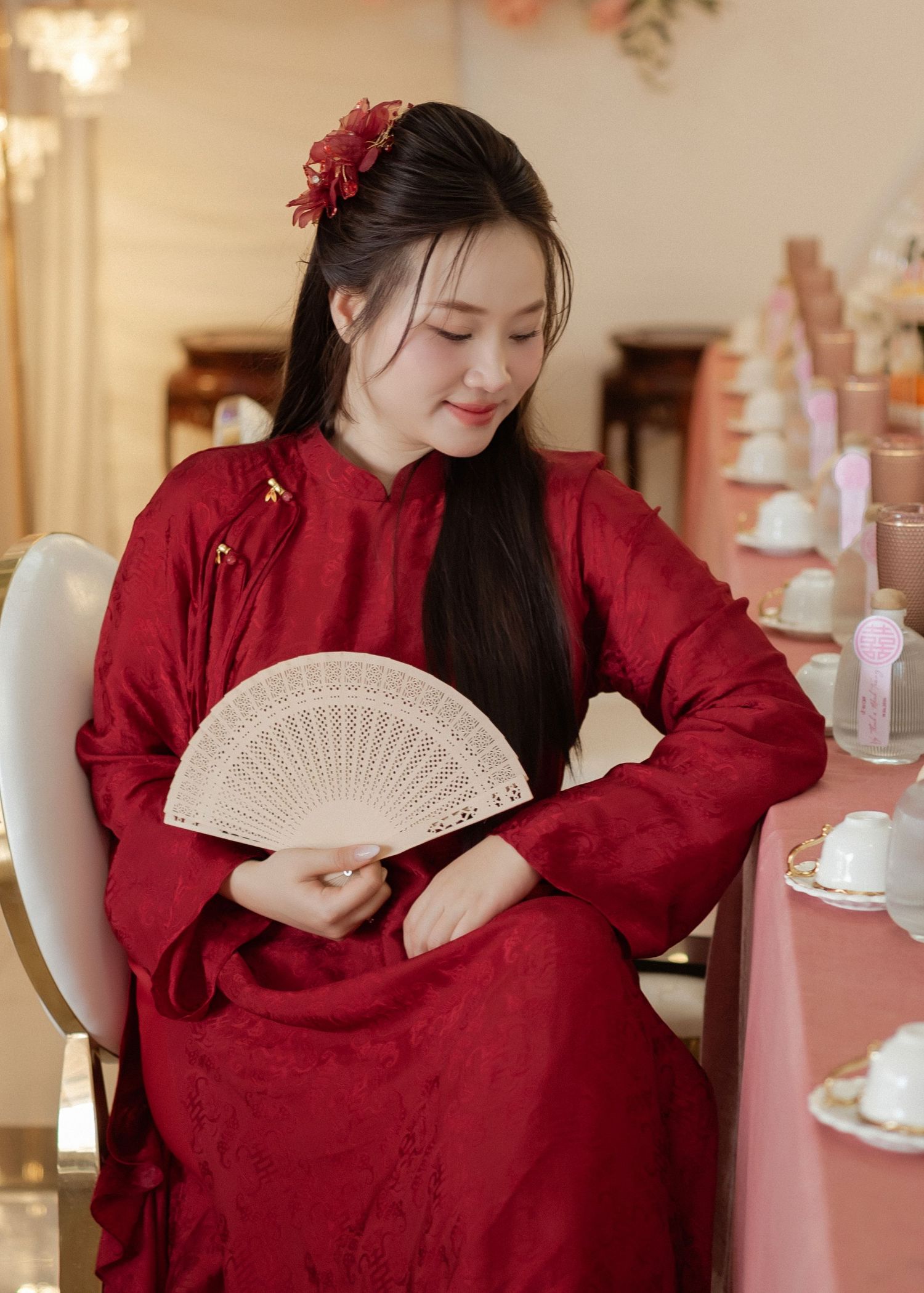 A woman in a traditional red Chinese dress holding a white folding fan sits at a pink table.