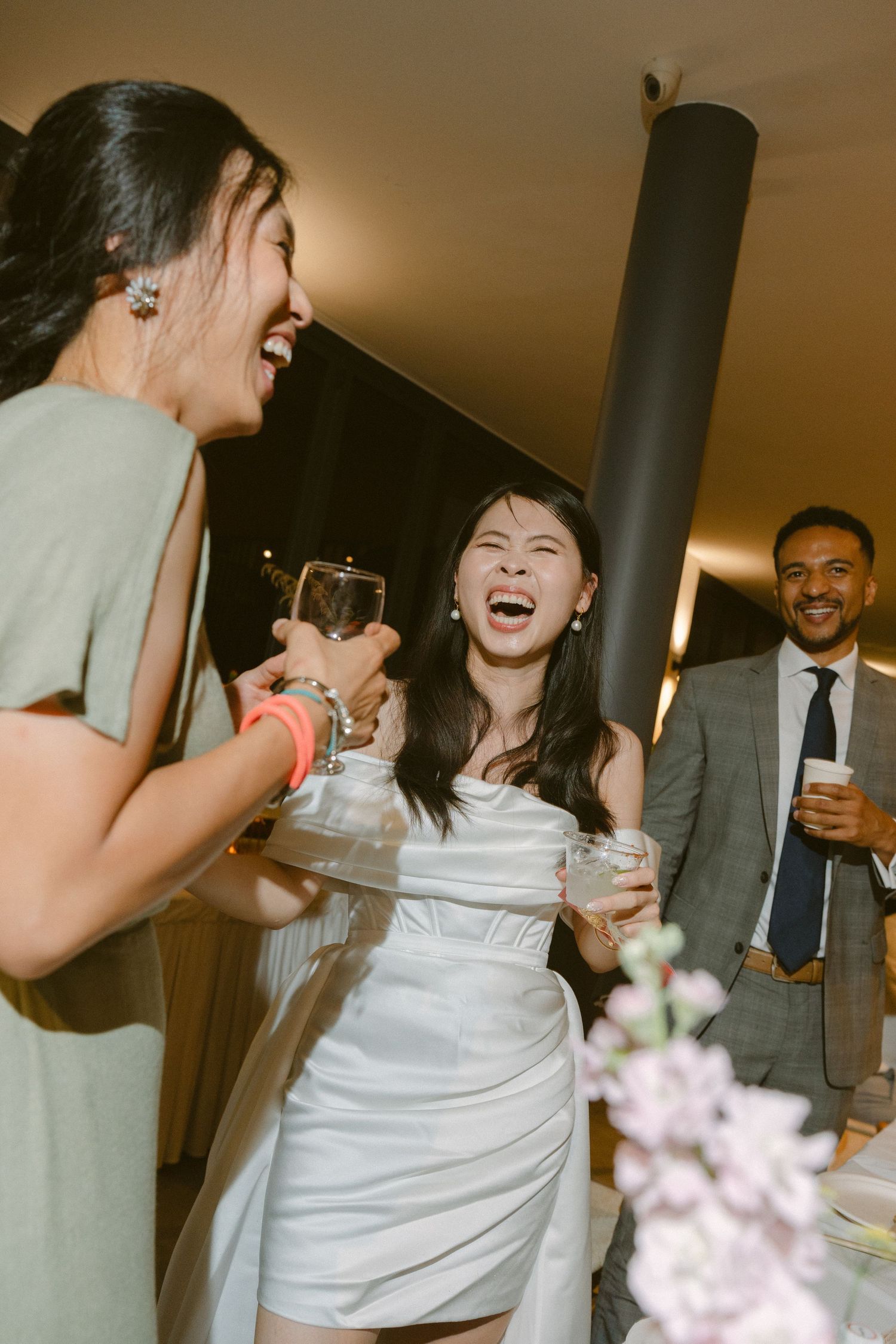 A joyful candid moment at a wedding reception shows people laughing together with soft lighting and floral decor.