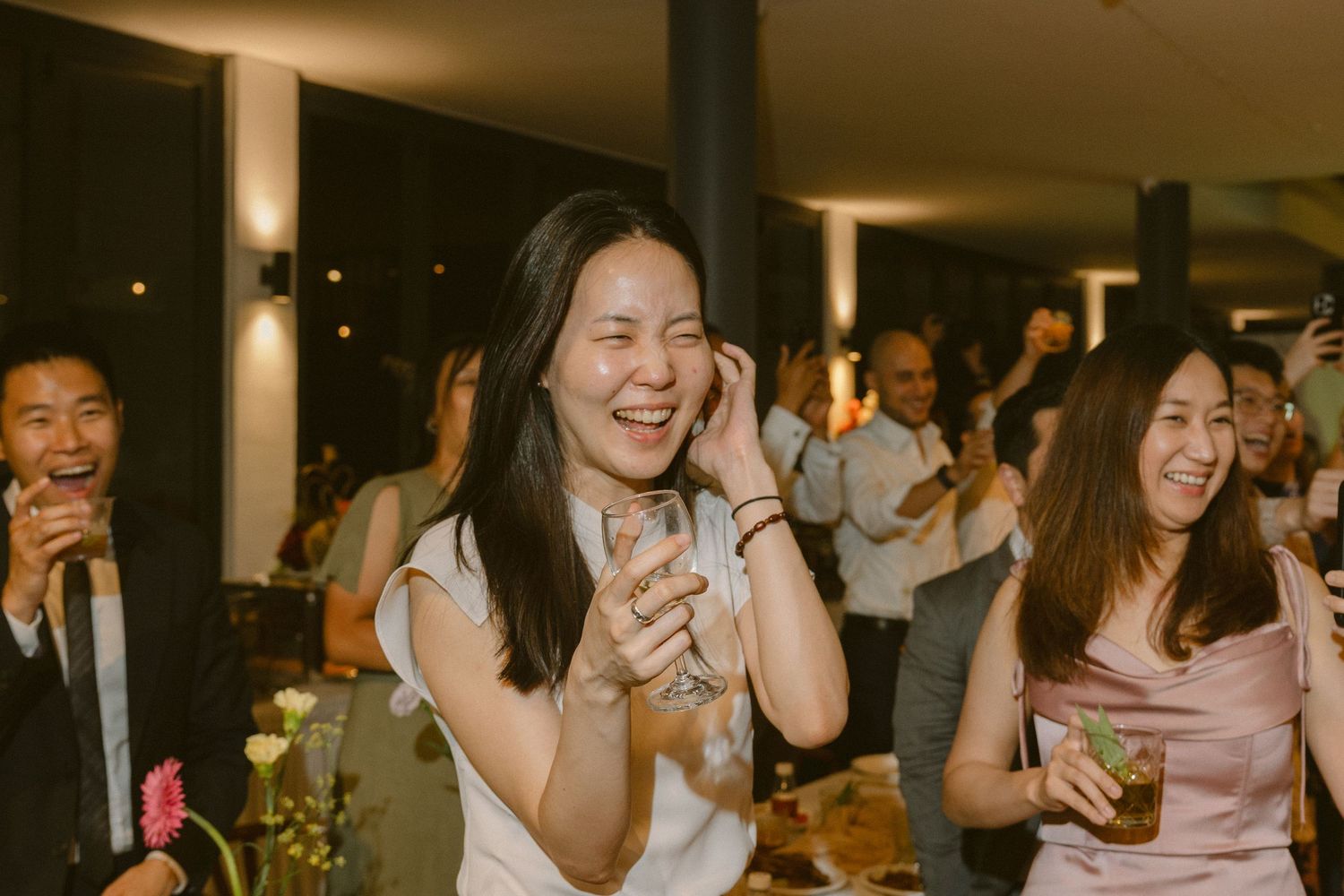 A group of friends laugh and smile together while enjoying drinks at a dimly lit indoor event.