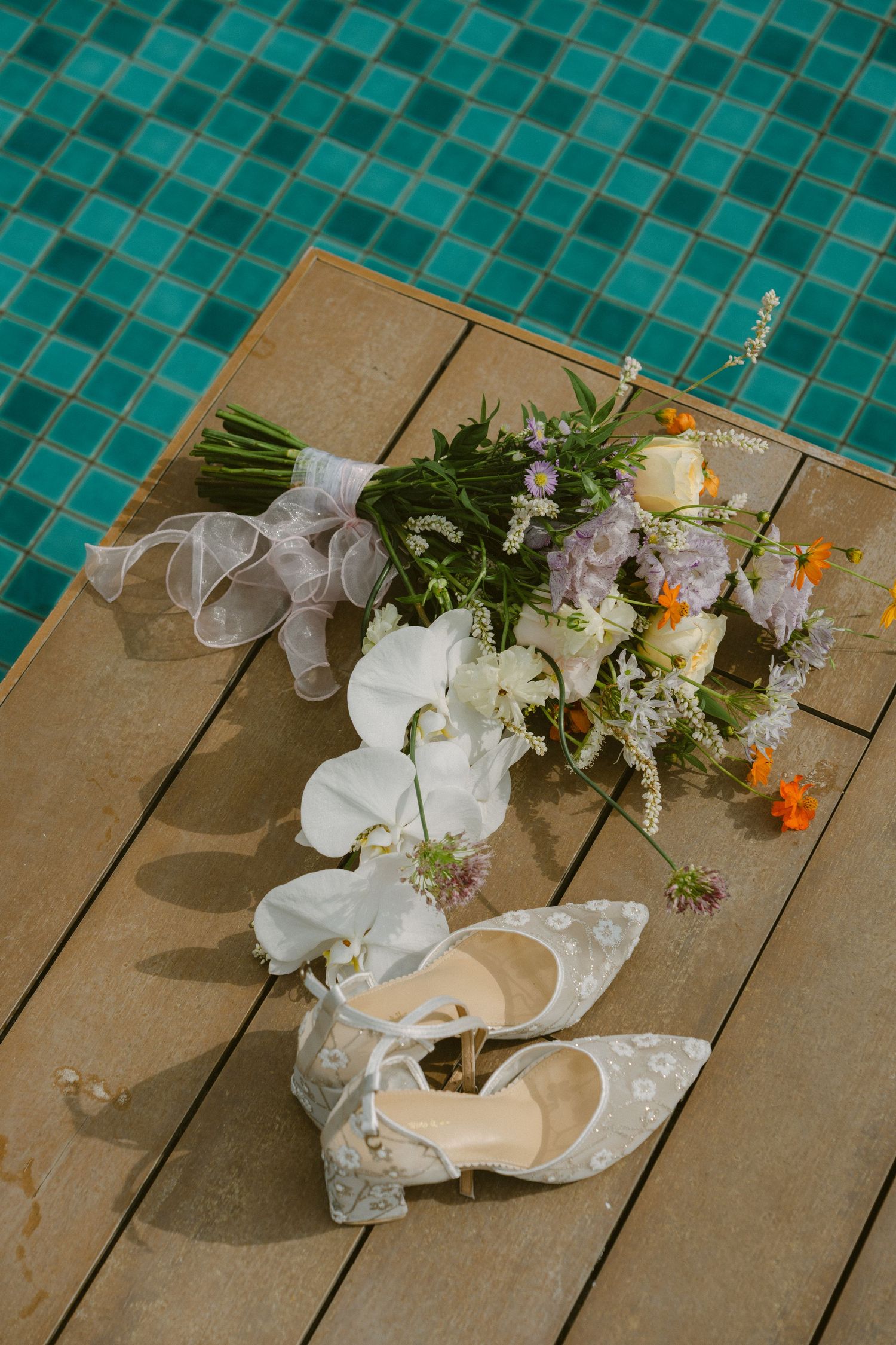 Wedding details featuring elegant white bridal shoes and a bouquet with orchids on wooden deck by turquoise tile pool.