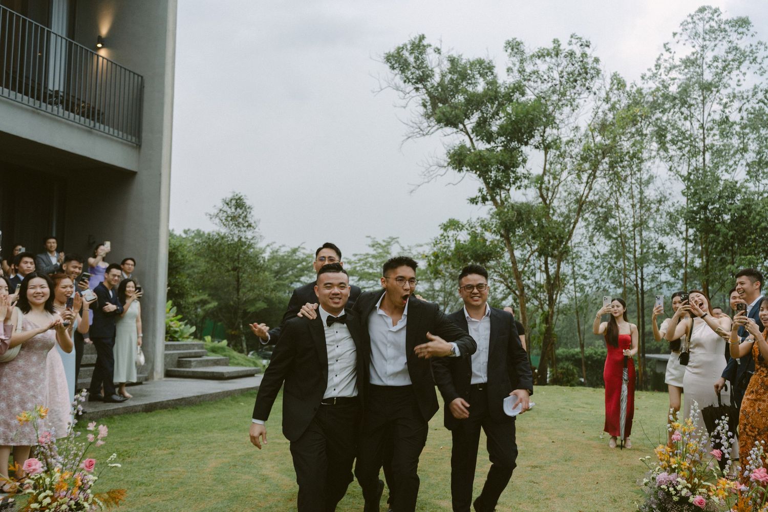 Wedding party members walk and pose together outdoors on a cloudy day amidst green landscaping.