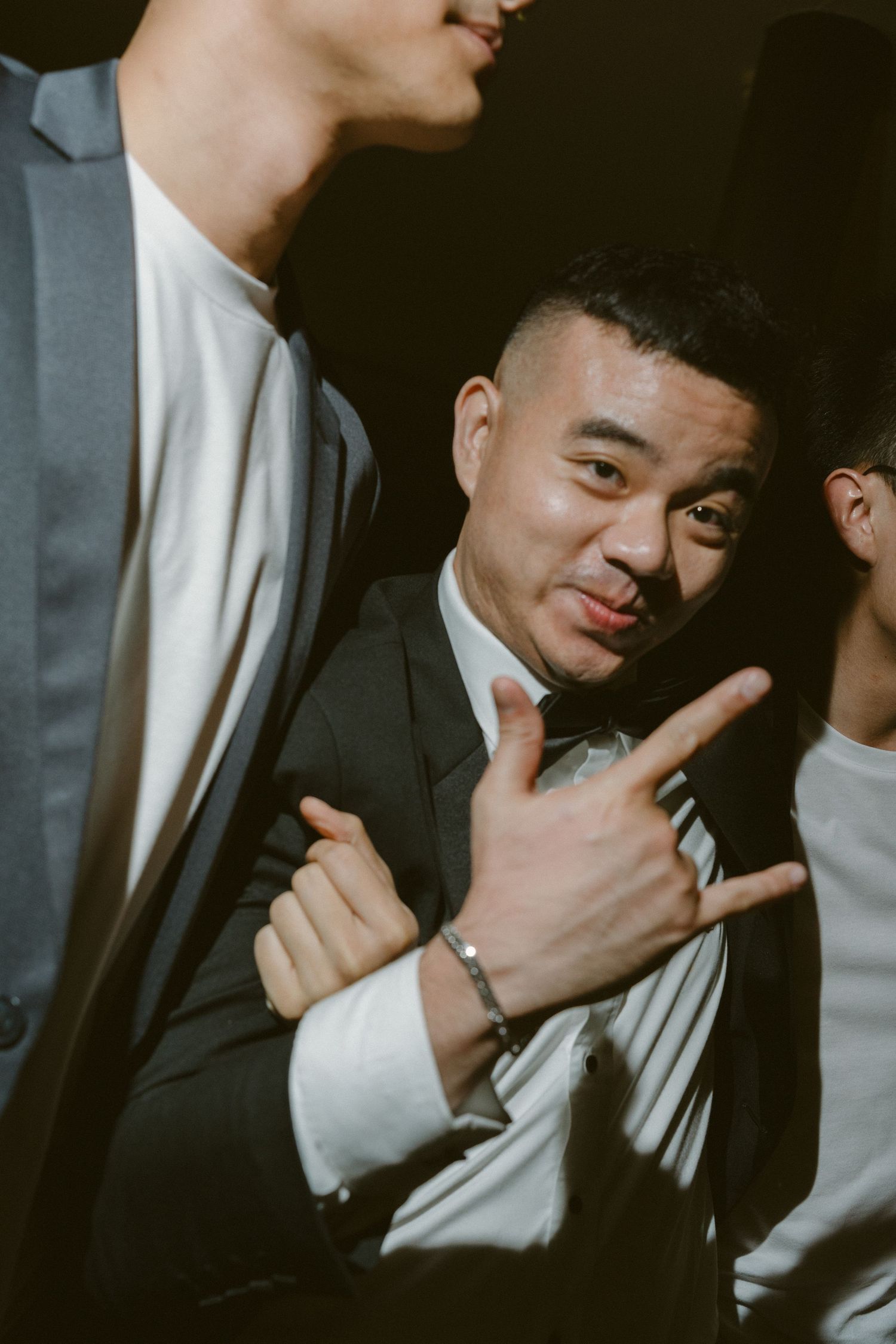 A person in a black suit making a hand gesture while others are partially visible in the frame.