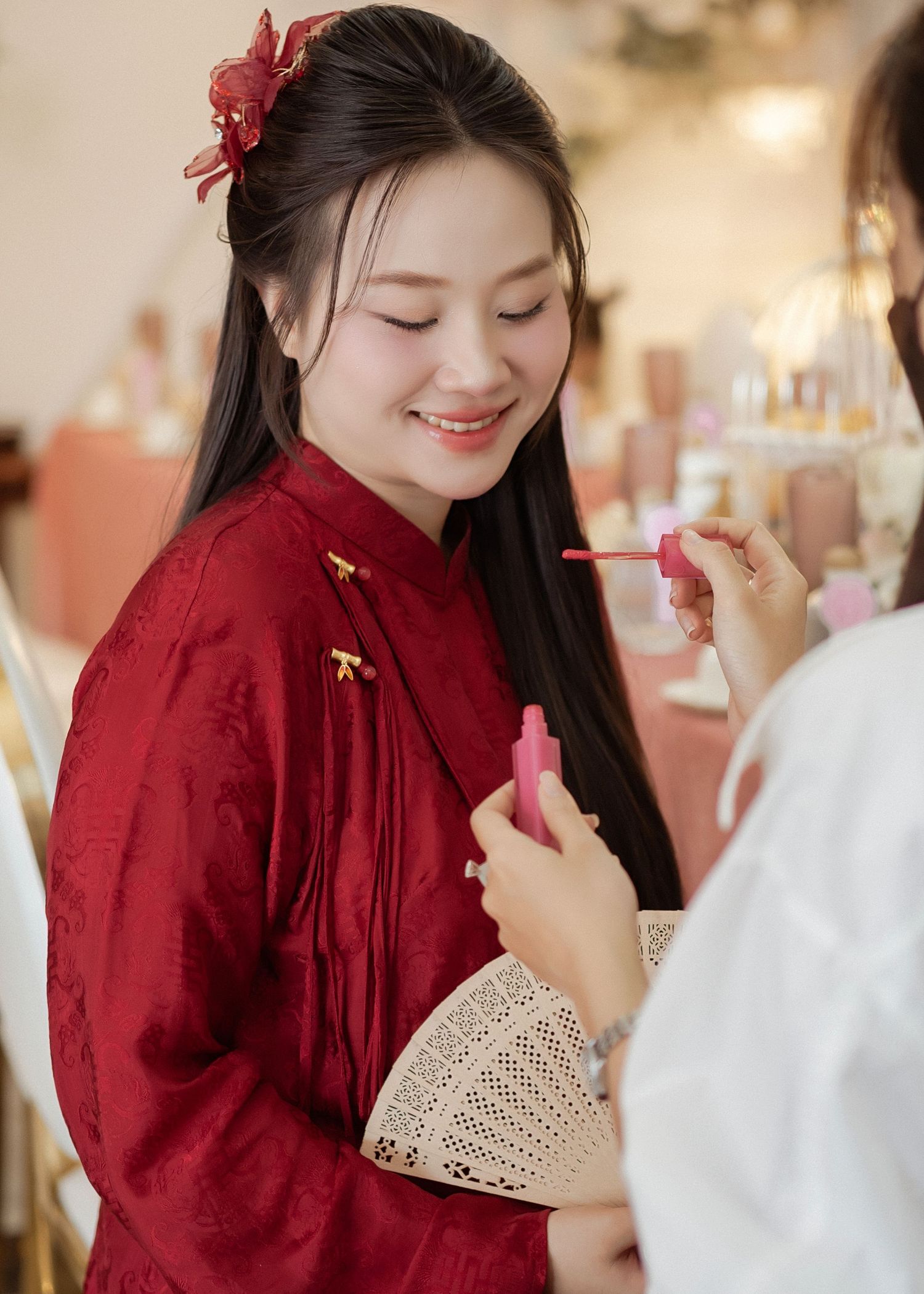 Person in traditional red Chinese qipao smiling while having makeup applied, with a red flower hair accessory.