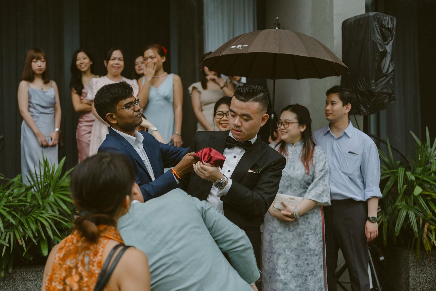 Wedding guests gather under a brown umbrella during an outdoor celebration at a lush garden venue.