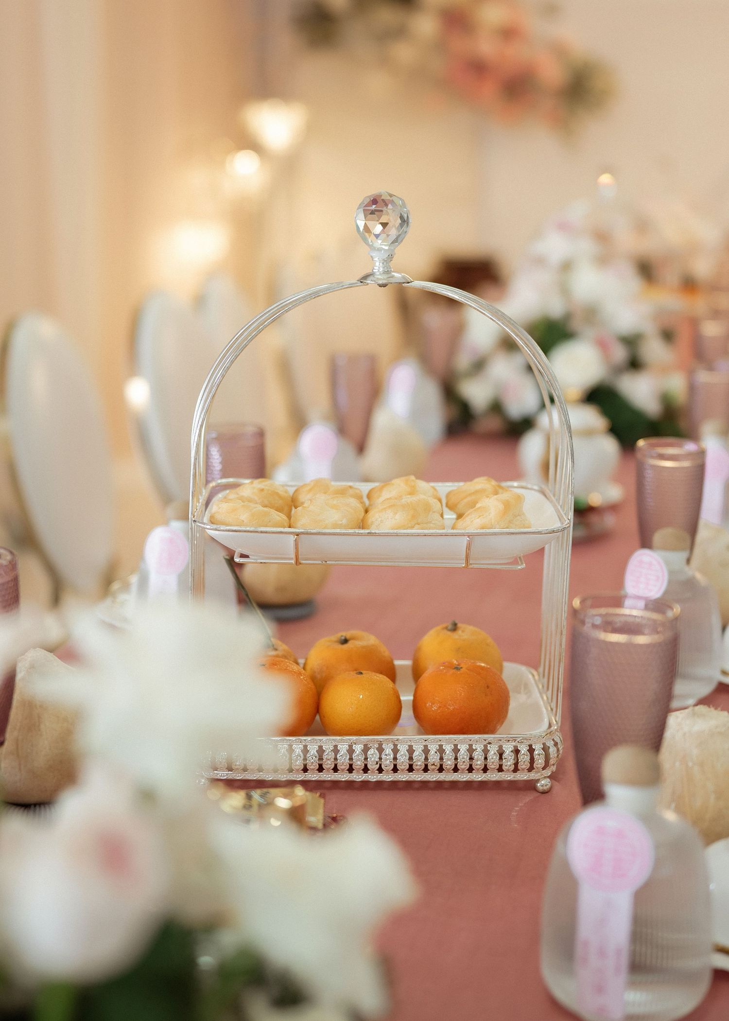 A tiered dessert stand displays fluffy pastries and oranges on a pink tablecloth with floral decor and drinks in the background.