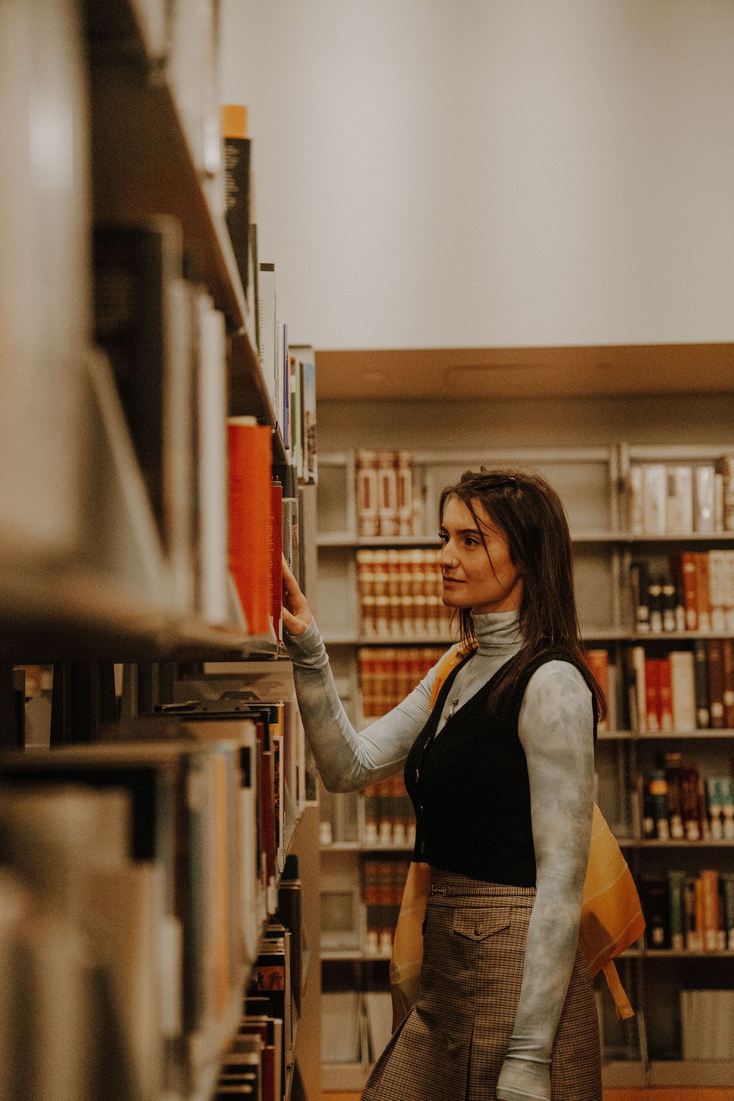 Person in turtleneck and dark vest searches for books in a cozy library setting with wooden shelves.