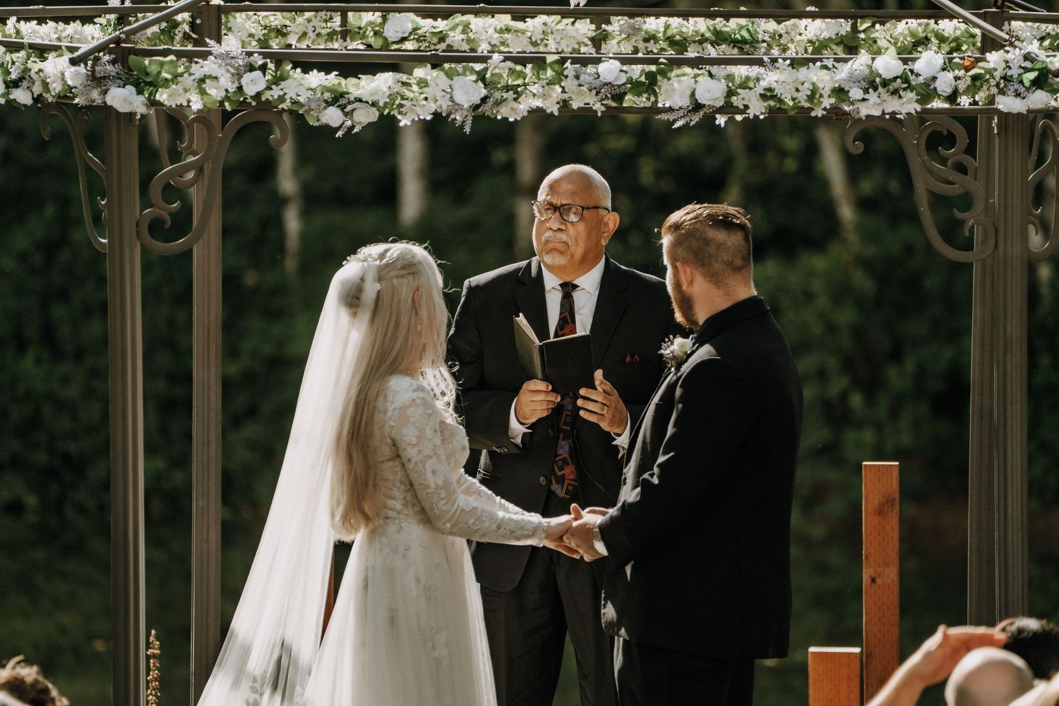 Wedding ceremony takes place under a floral arch with an officiant conducting the service outdoors.