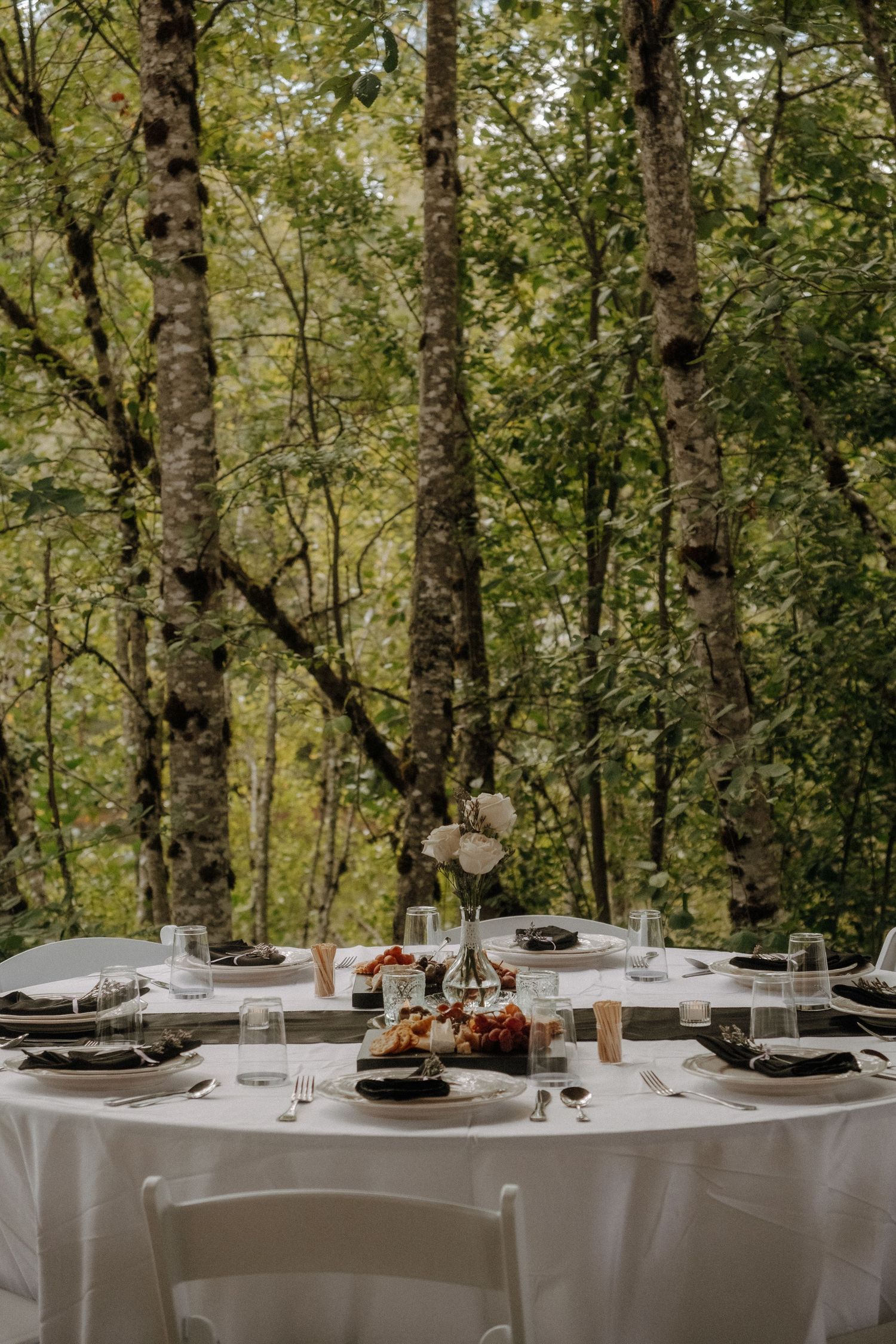 Elegant wedding reception table set in a forest with white linens and rustic decorations.