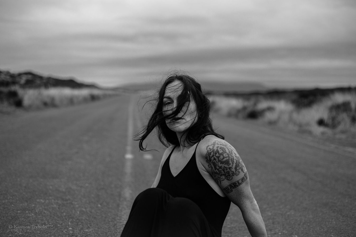 Black and white portrait of person in dress with tattoo on shoulder on empty country road.