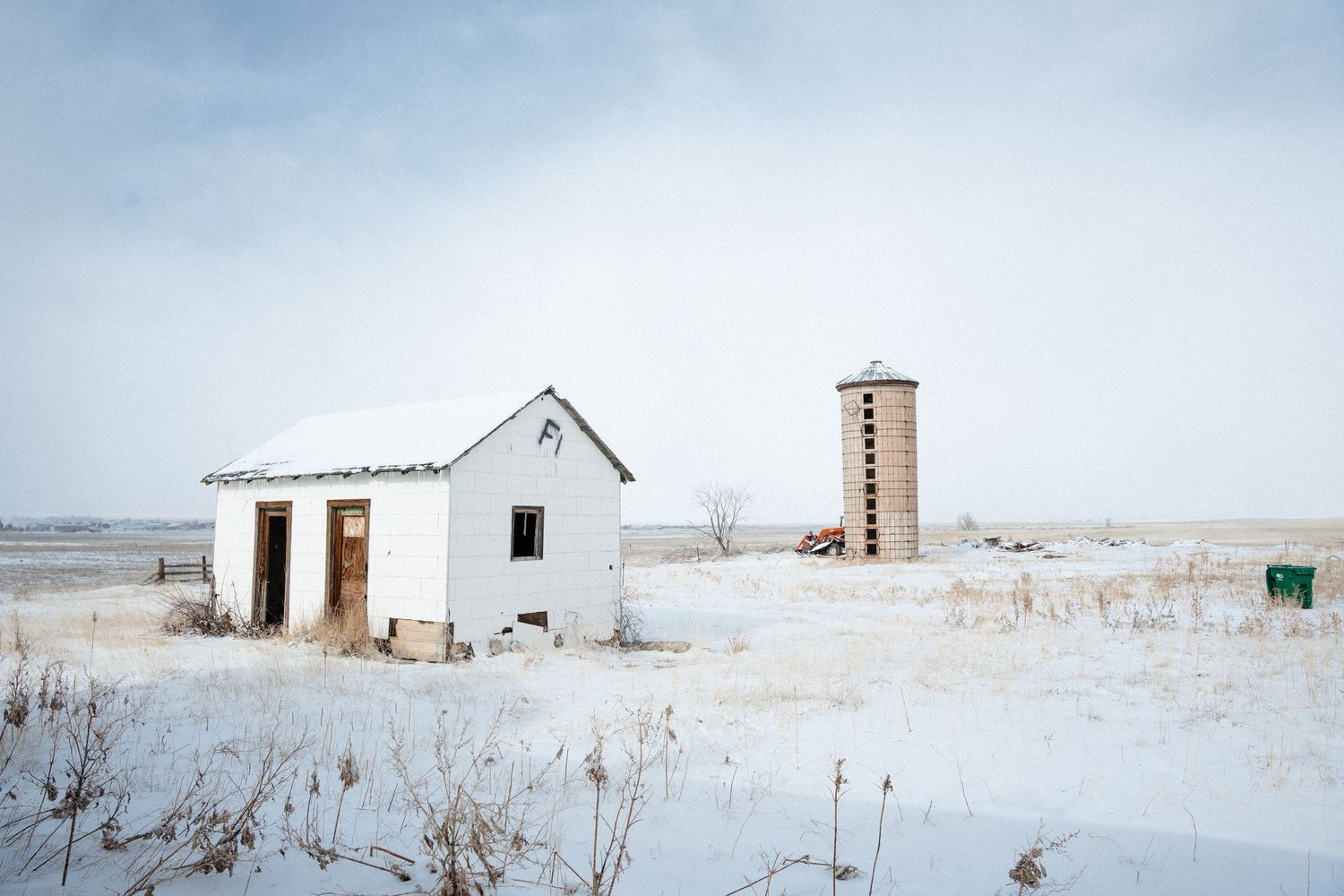 A small white abandoned building and water tower stand isolated in a snowy winter landscape on the prairie.