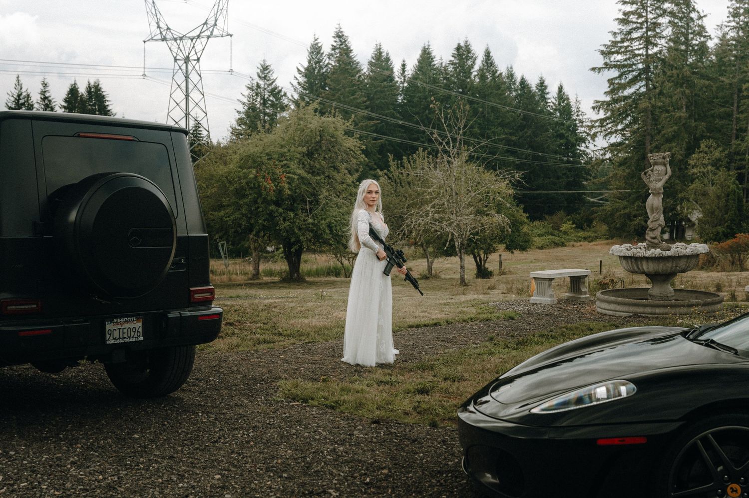 A elegant white dress contrasts against evergreen trees and parked vehicles on a rural property.