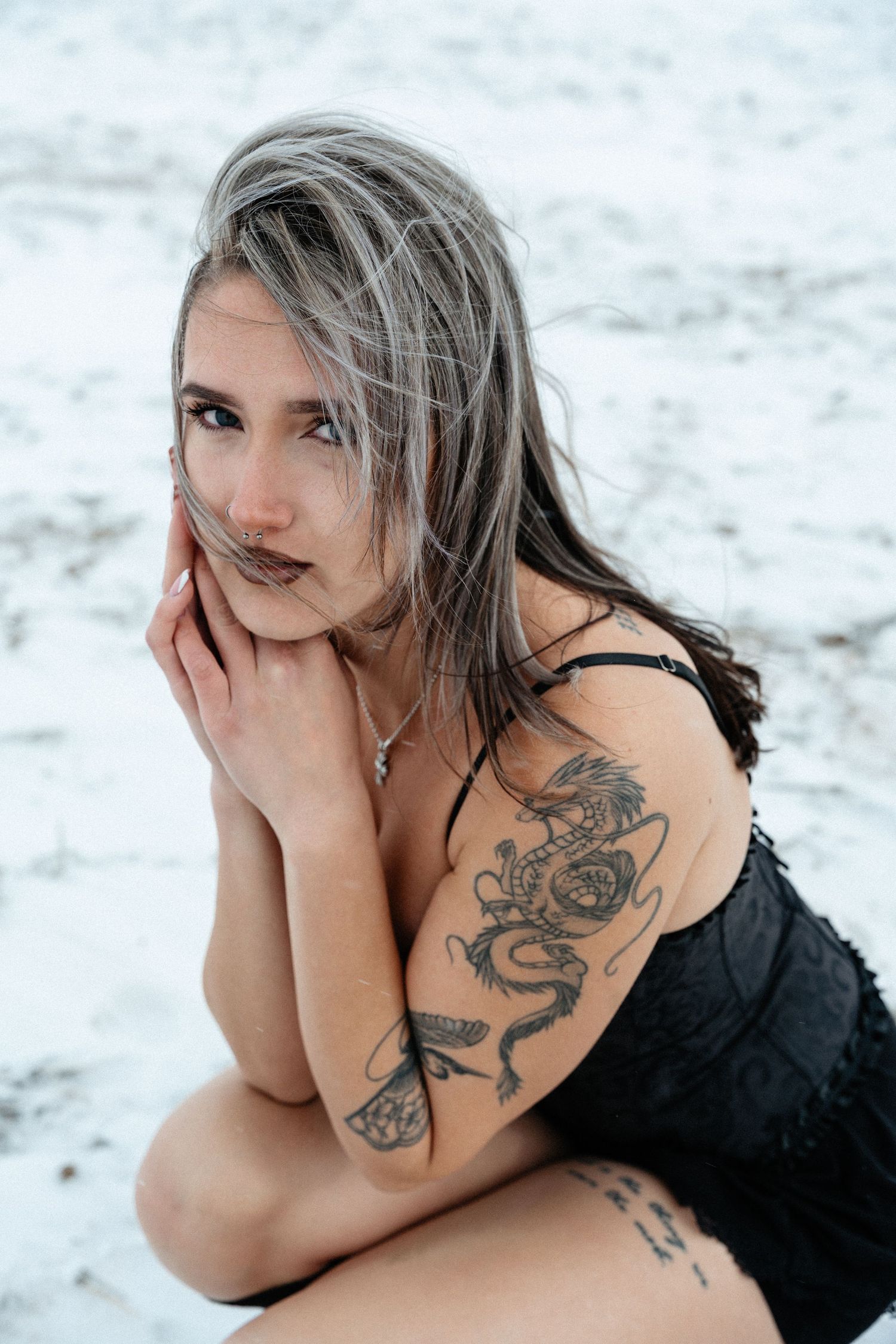 Model with long hair poses on white sand beach showing dragon tattoo design on arm.