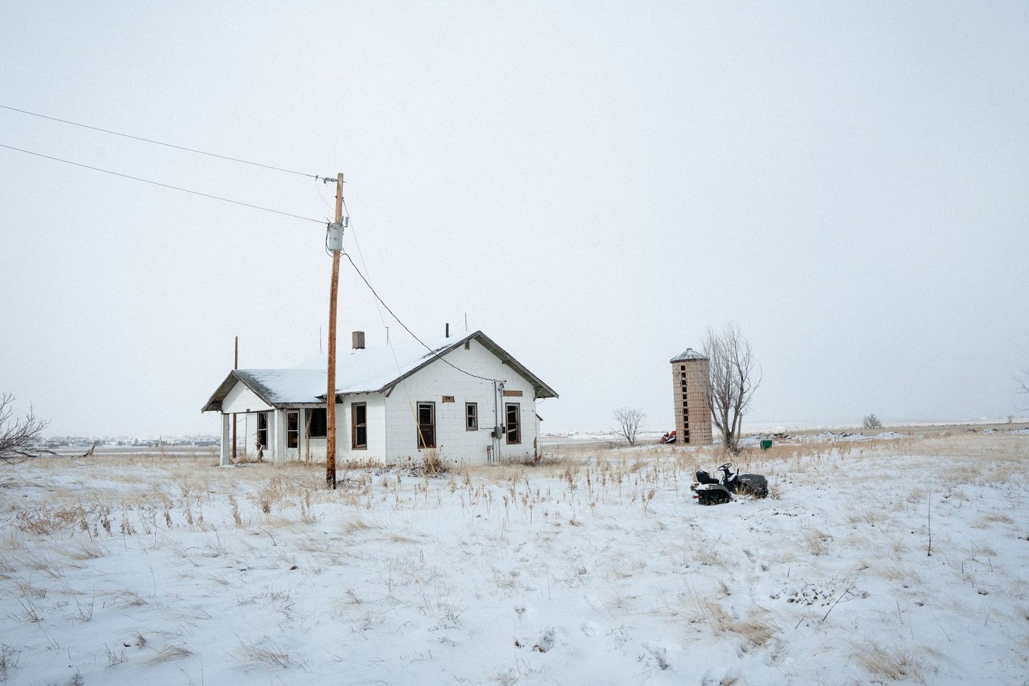 An abandoned white farmhouse and silo stand isolated in a snowy winter landscape with utility poles nearby.