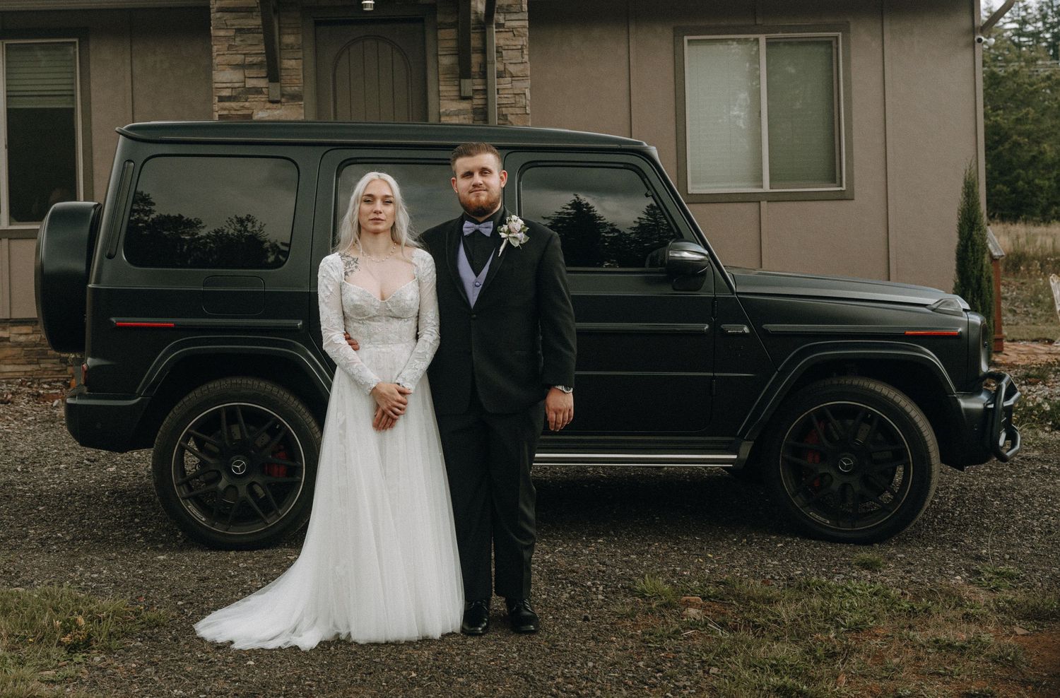 Well-dressed couple poses in formal attire next to black Mercedes G-Wagon outside stone building.