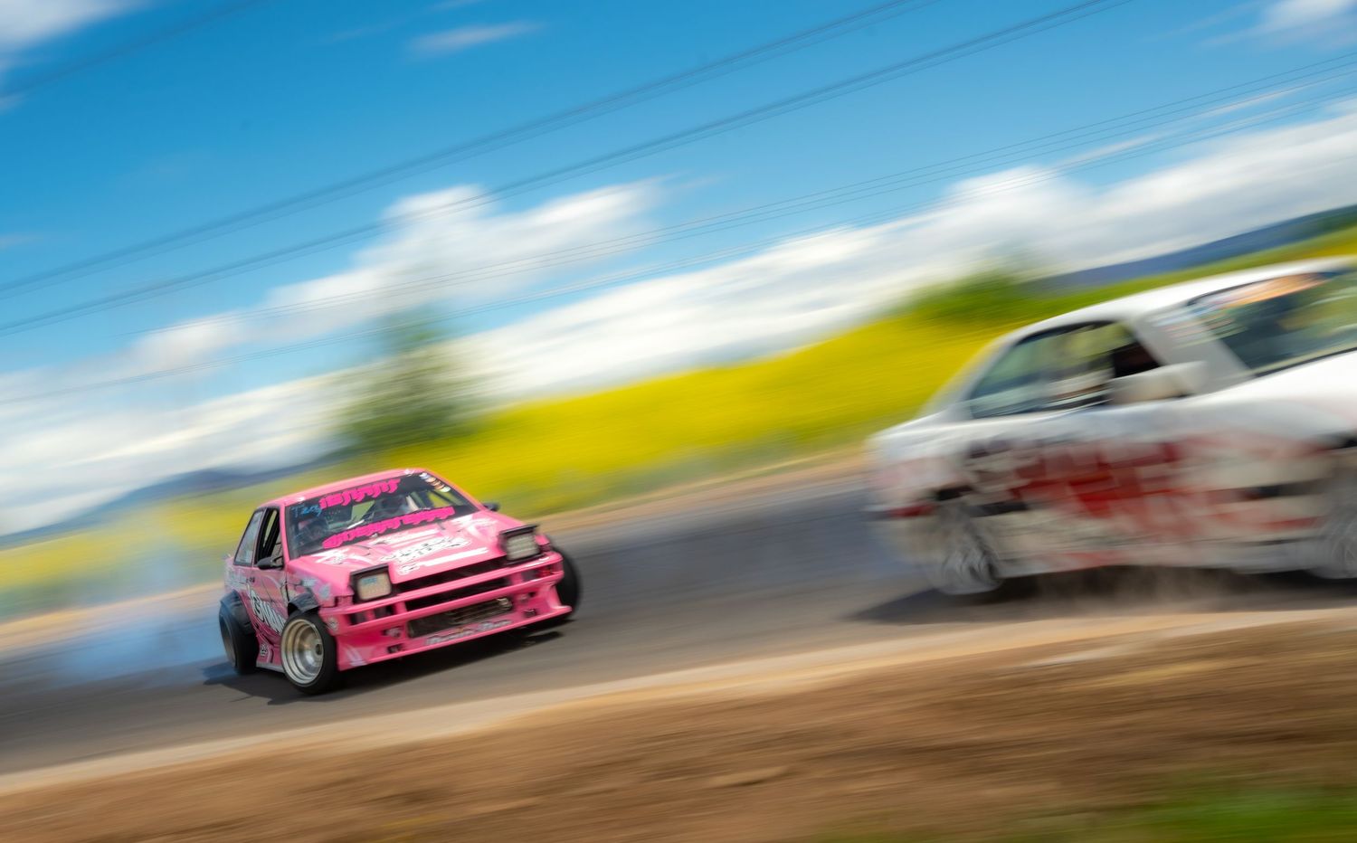 Racing cars zoom around a racetrack creating motion blur against a vibrant blue sky and green landscape.