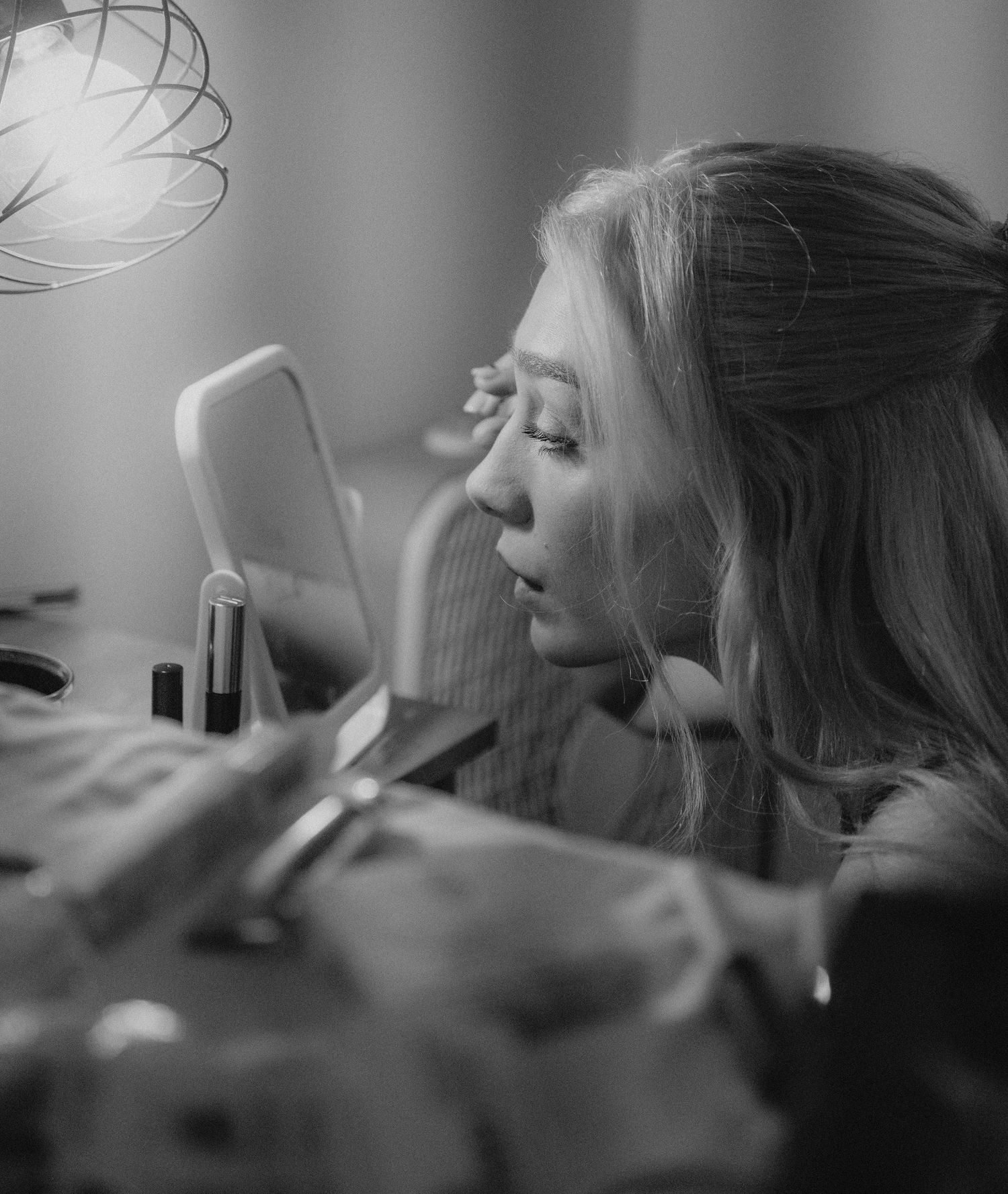 Black and white photo of a person lying down looking at their mobile phone in low light, with makeup products nearby.
