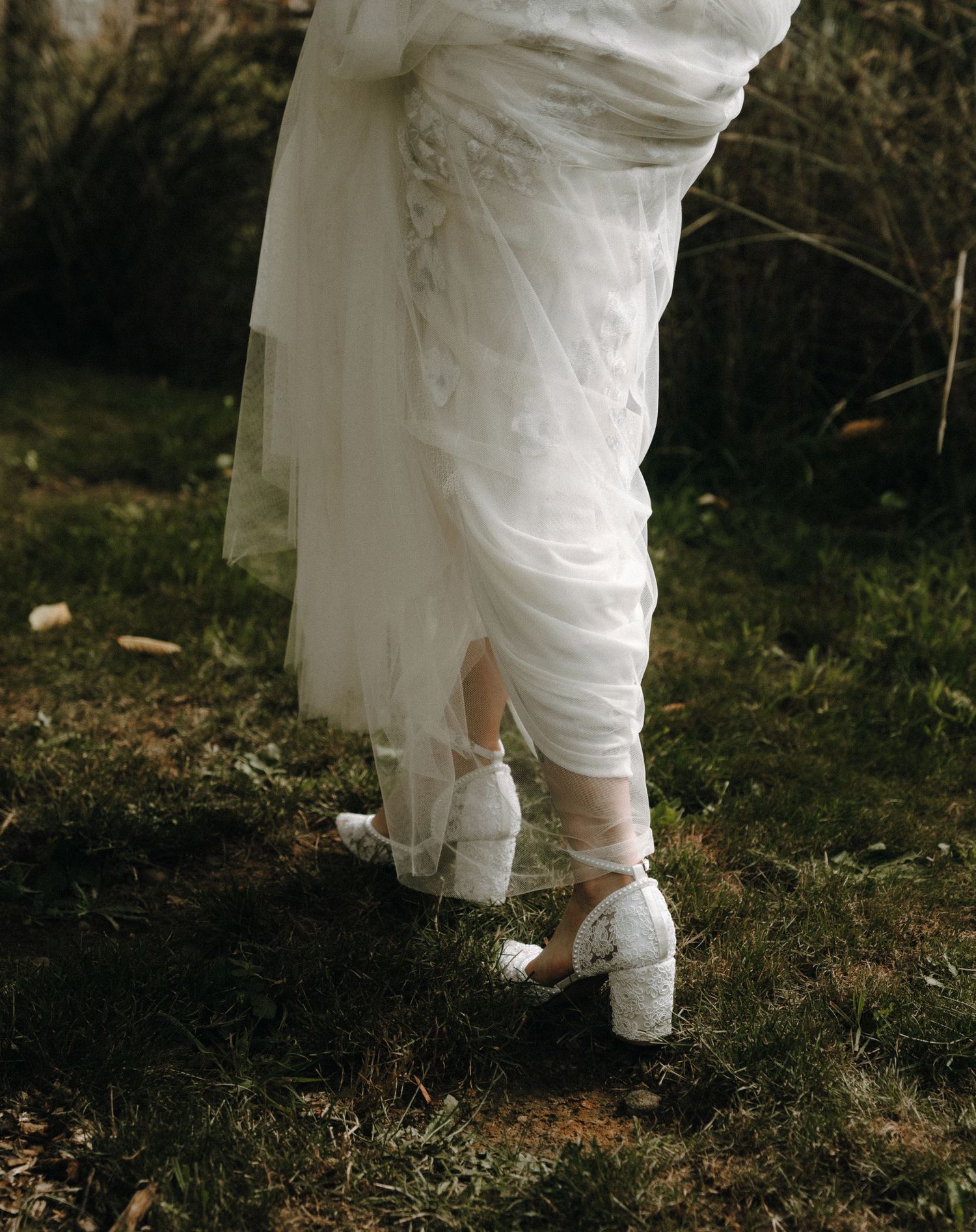 White wedding dress hem and vintage-inspired block heel shoes shown against grass in a romantic outdoor setting.