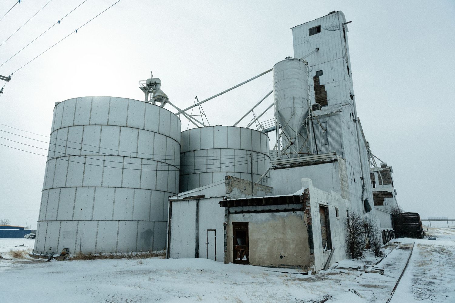 Large industrial grain storage facility with white metal silos and elevator tower covered in snow during winter.