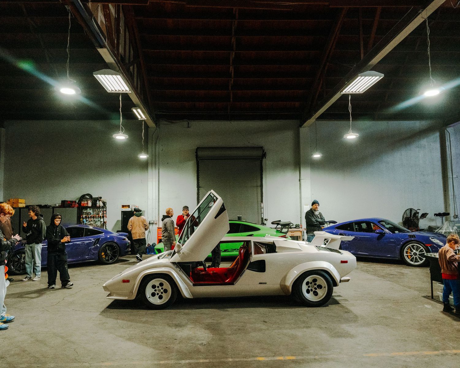 An underground parking garage displays a collection of luxury and exotic sports cars lined up under fluorescent lighting.