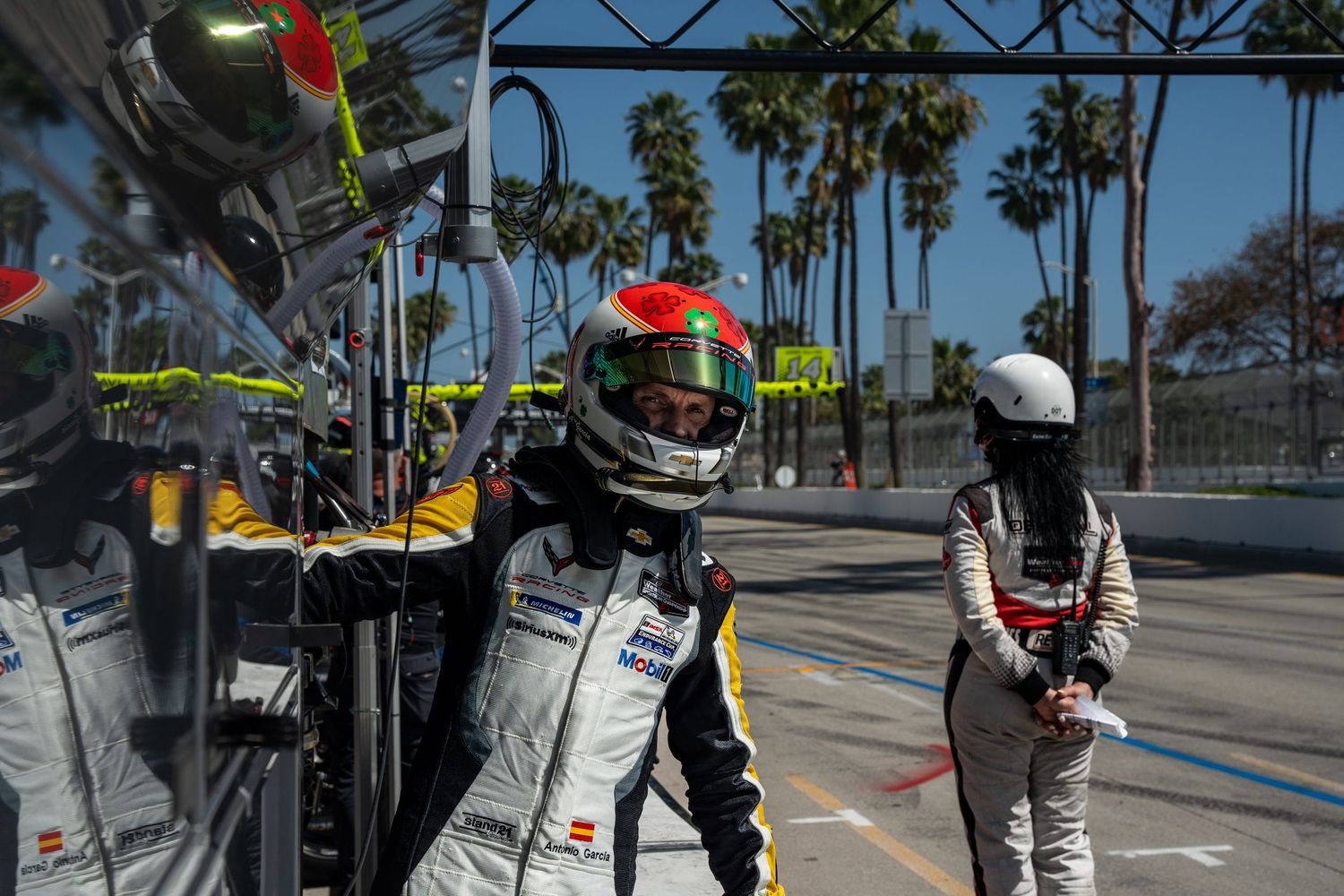 Drivers in race suits walking through pit lane with palm trees in the background at a sunny race track.