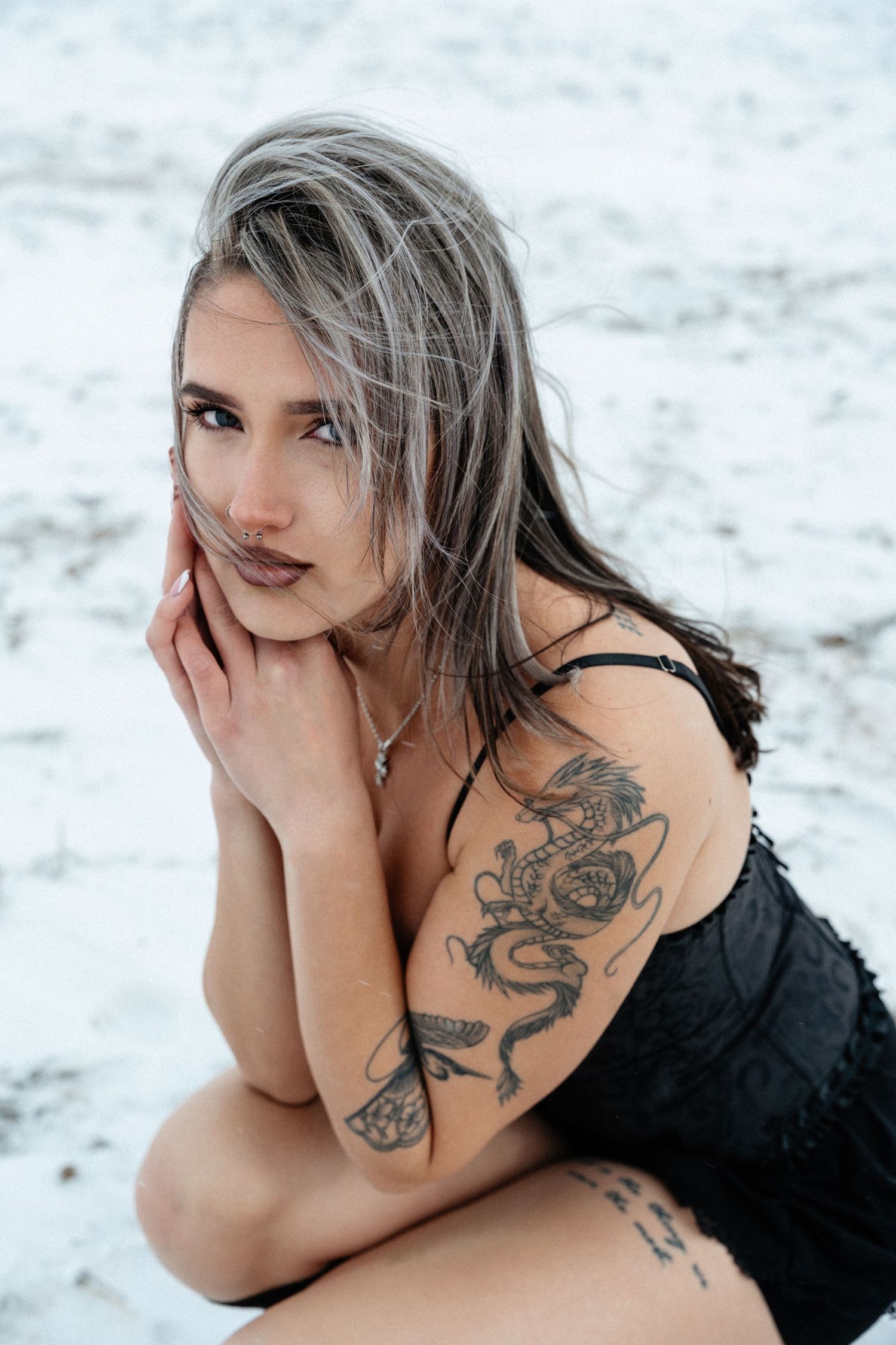 Woman with gray hair and tattoos poses on white snow wearing black dress in artistic portrait series.