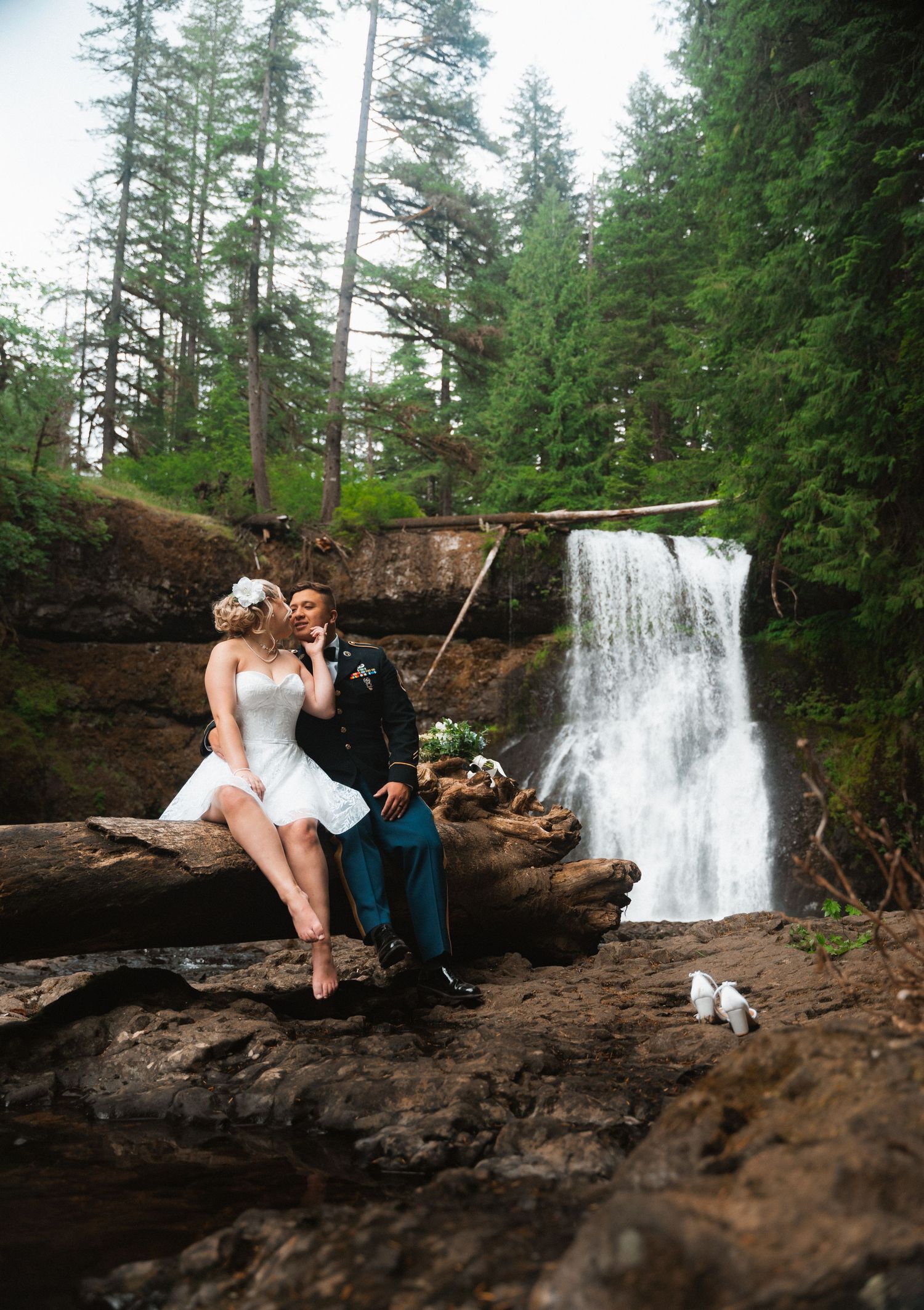 Wedding couple sits on rocky cliff with scenic waterfall cascading behind them in lush forest setting.