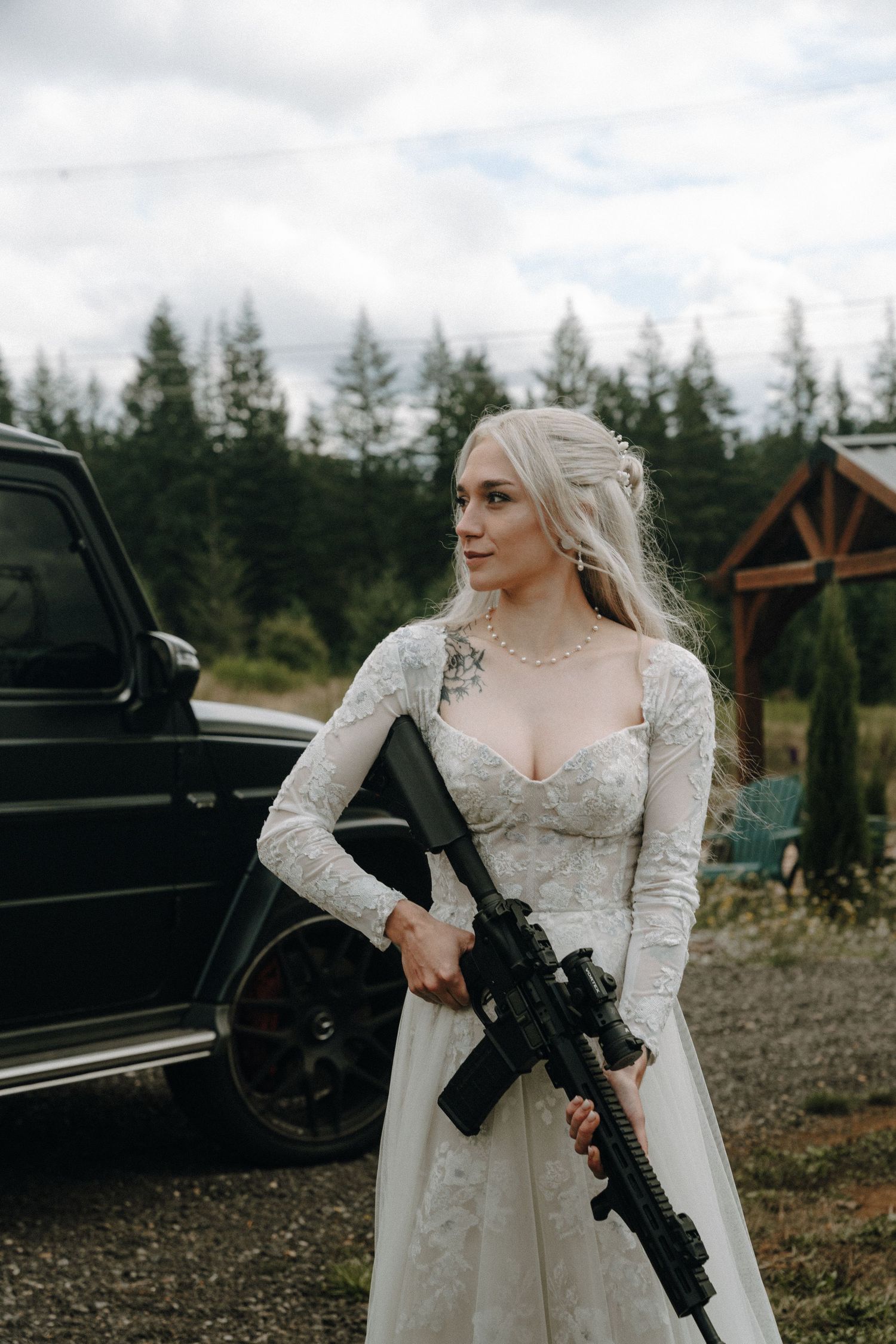A bride in a white lace wedding dress holds a black tactical rifle while standing next to a vehicle in a forest setting.