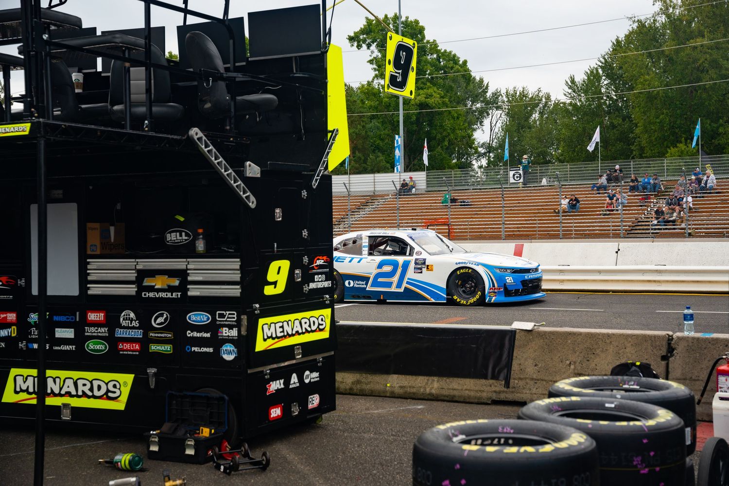 A NASCAR race car slides off the track near pit row with stacked tires and equipment in the foreground.