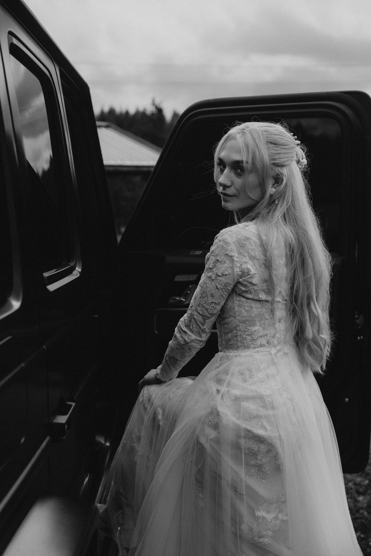 A bride in a flowing white dress and long hair enters a black vintage car in a moody black and white composition.