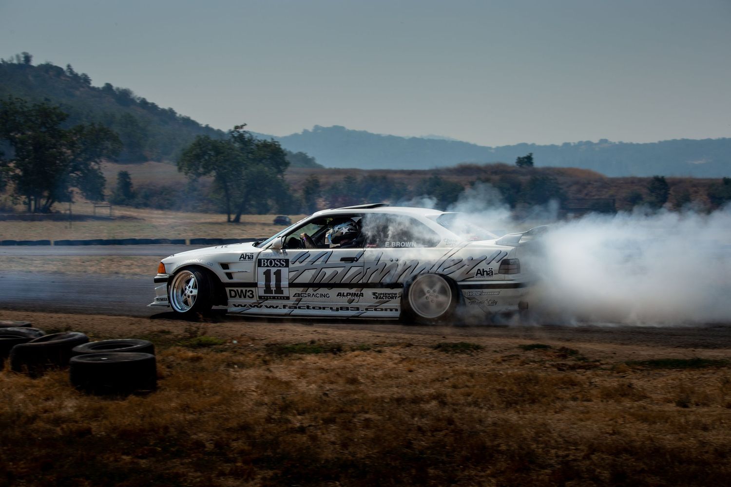 A silver race car performing a drift maneuver on a dirt track with tire smoke billowing behind it.