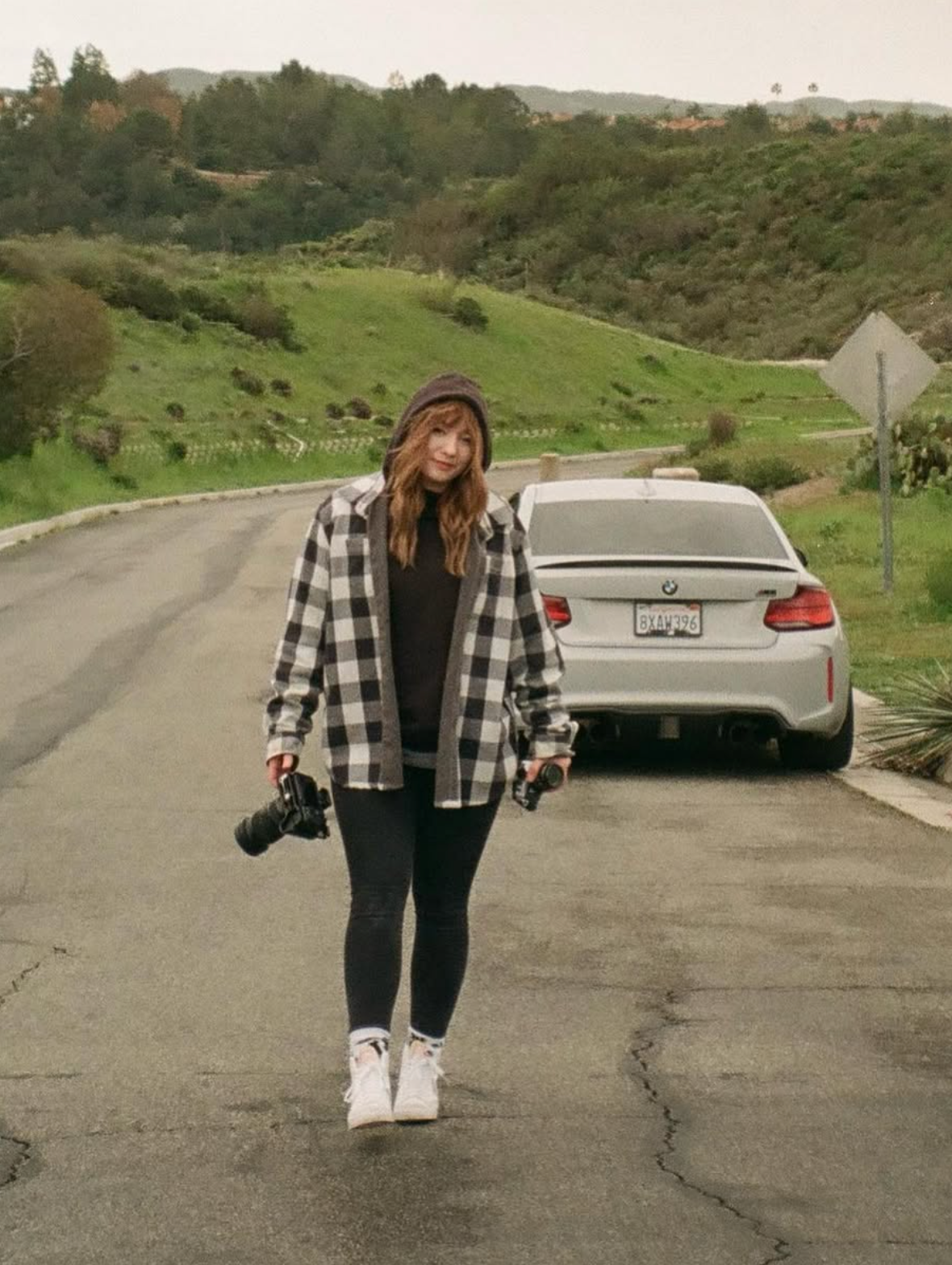Photographer in plaid flannel and black leggings standing on winding rural road with car parked in background.