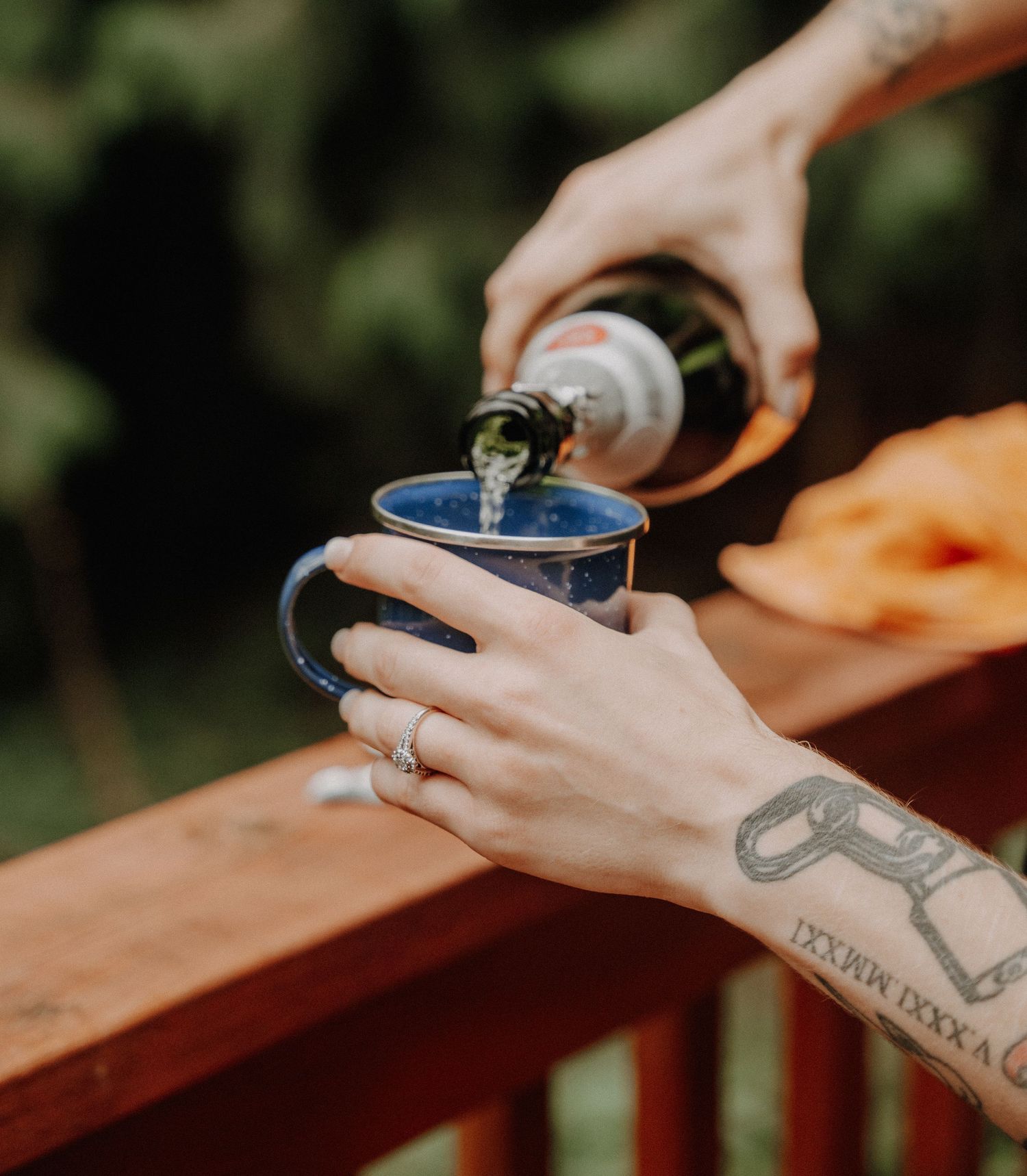 Someone with a tattoo pours a drink into a blue enamel camping mug while sitting at an outdoor wooden railing.