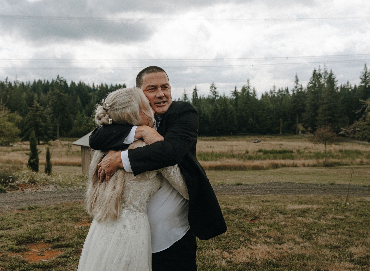 A couple embraces and dances together in an open field with trees and mountains in the background.