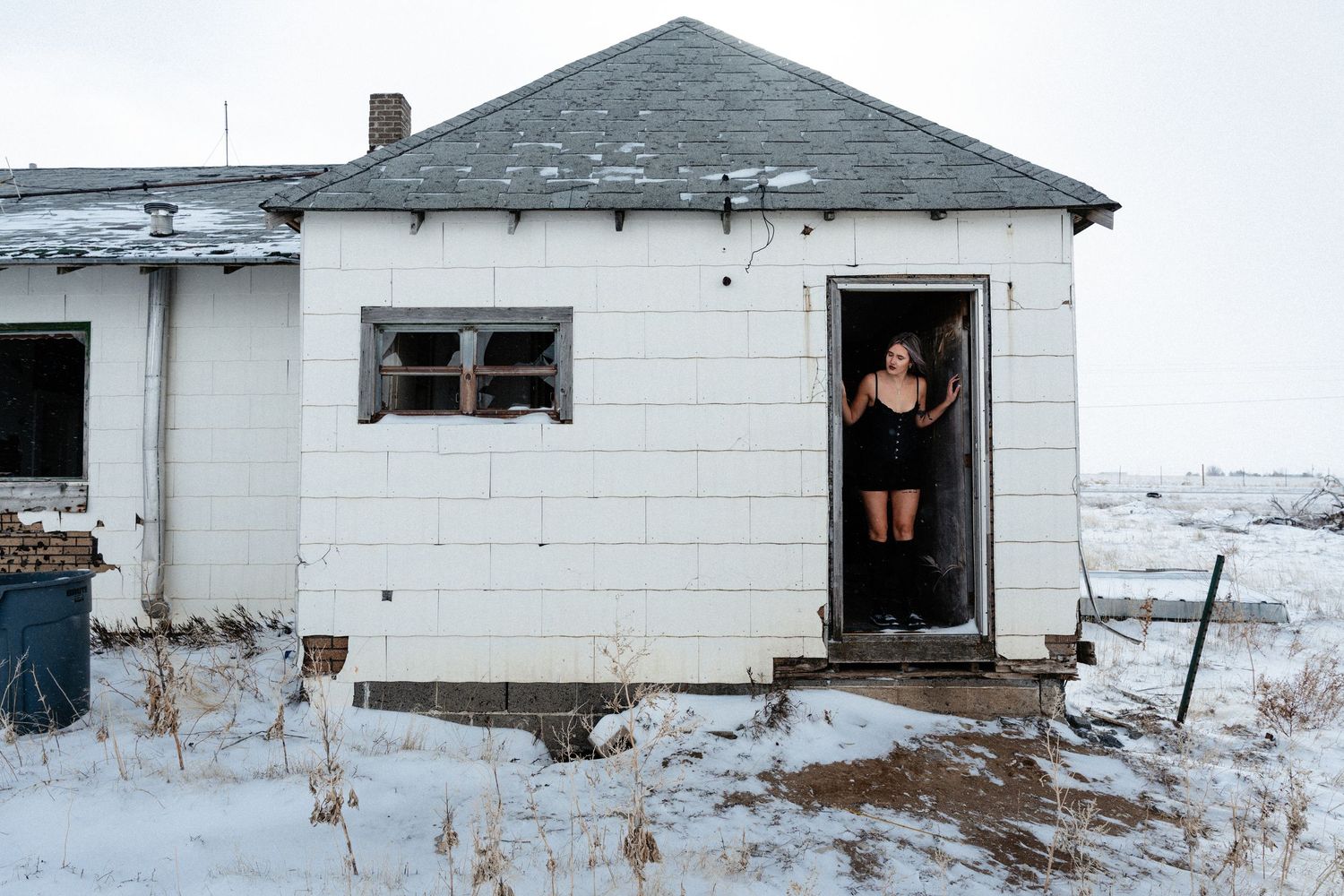 A dilapidated white house with multiple doorways stands isolated in a snowy rural landscape.