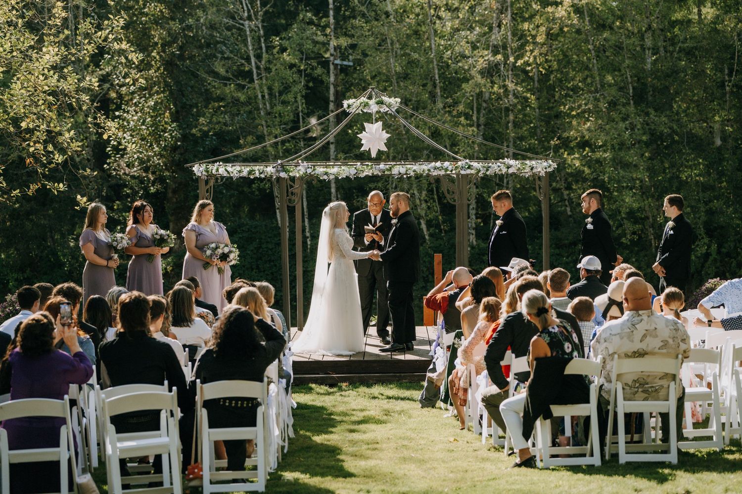 Outdoor wedding ceremony in a wooded setting with guests seated in white chairs and a decorated gazebo altar.