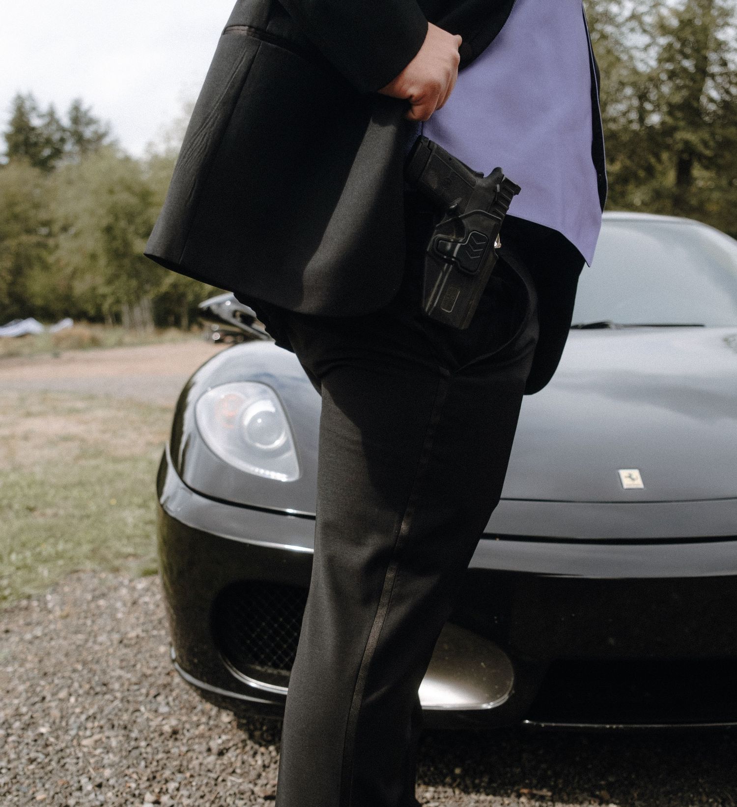 A person in black clothing stands next to a dark luxury sports car, with a holstered firearm visible.