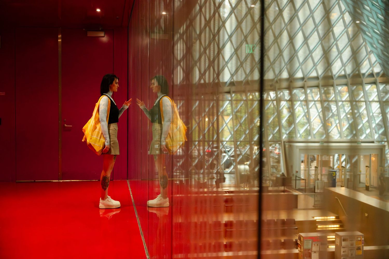Two figures with yellow bags stand in a modern hallway with red walls and geometric glass windows.