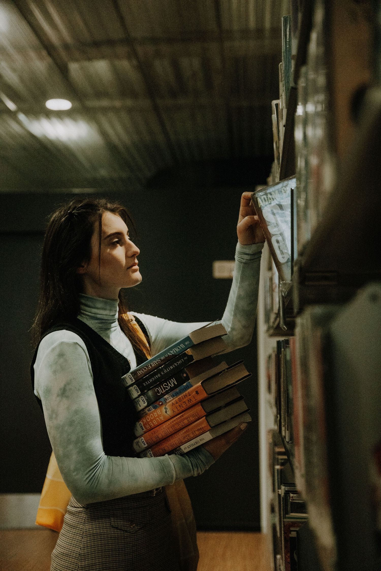 Woman in turtleneck browses library shelves while carrying stack of vintage books in dim lighting.