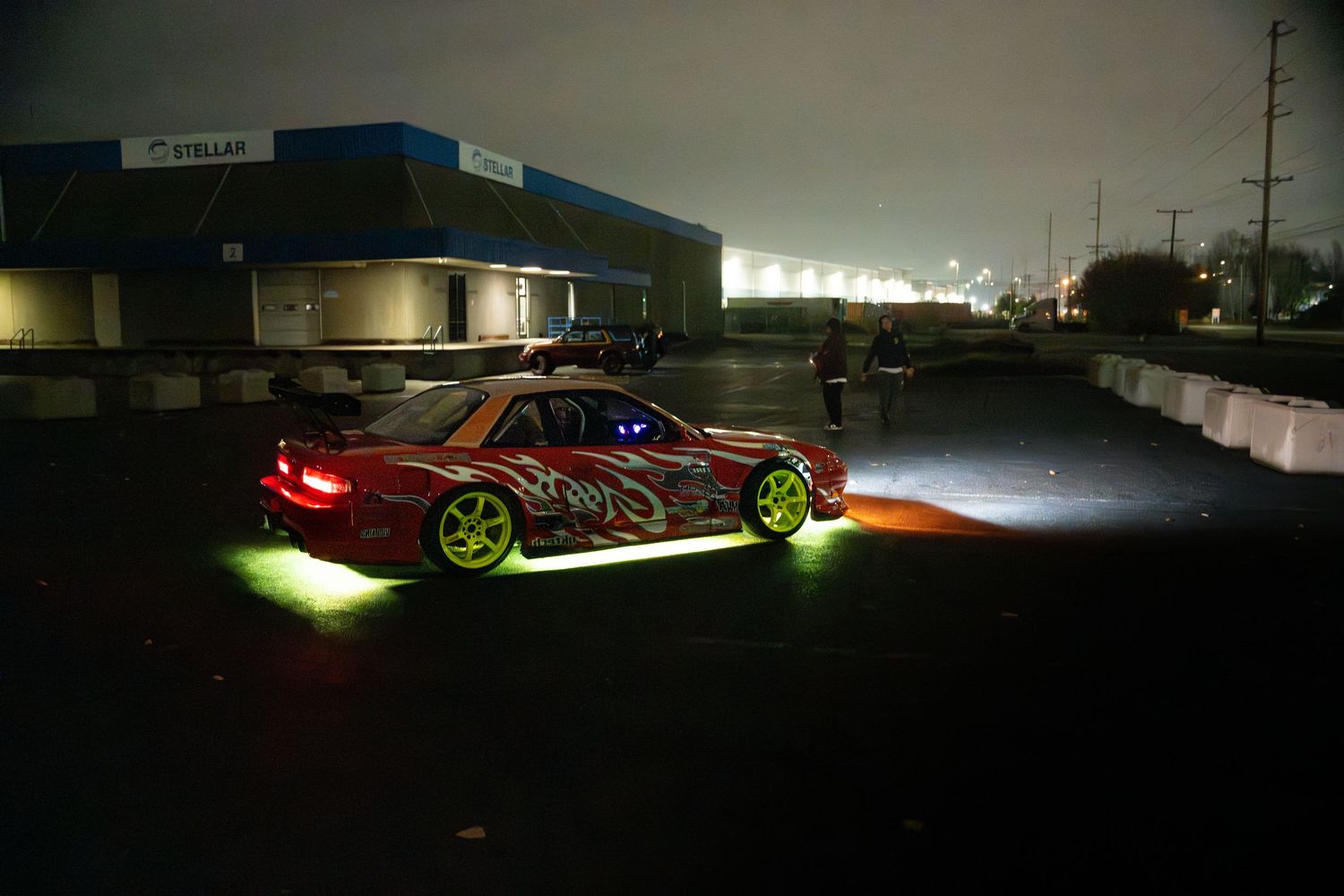 Modified sports car with neon underglow lights parked in dark parking lot at night with illuminated buildings in background.