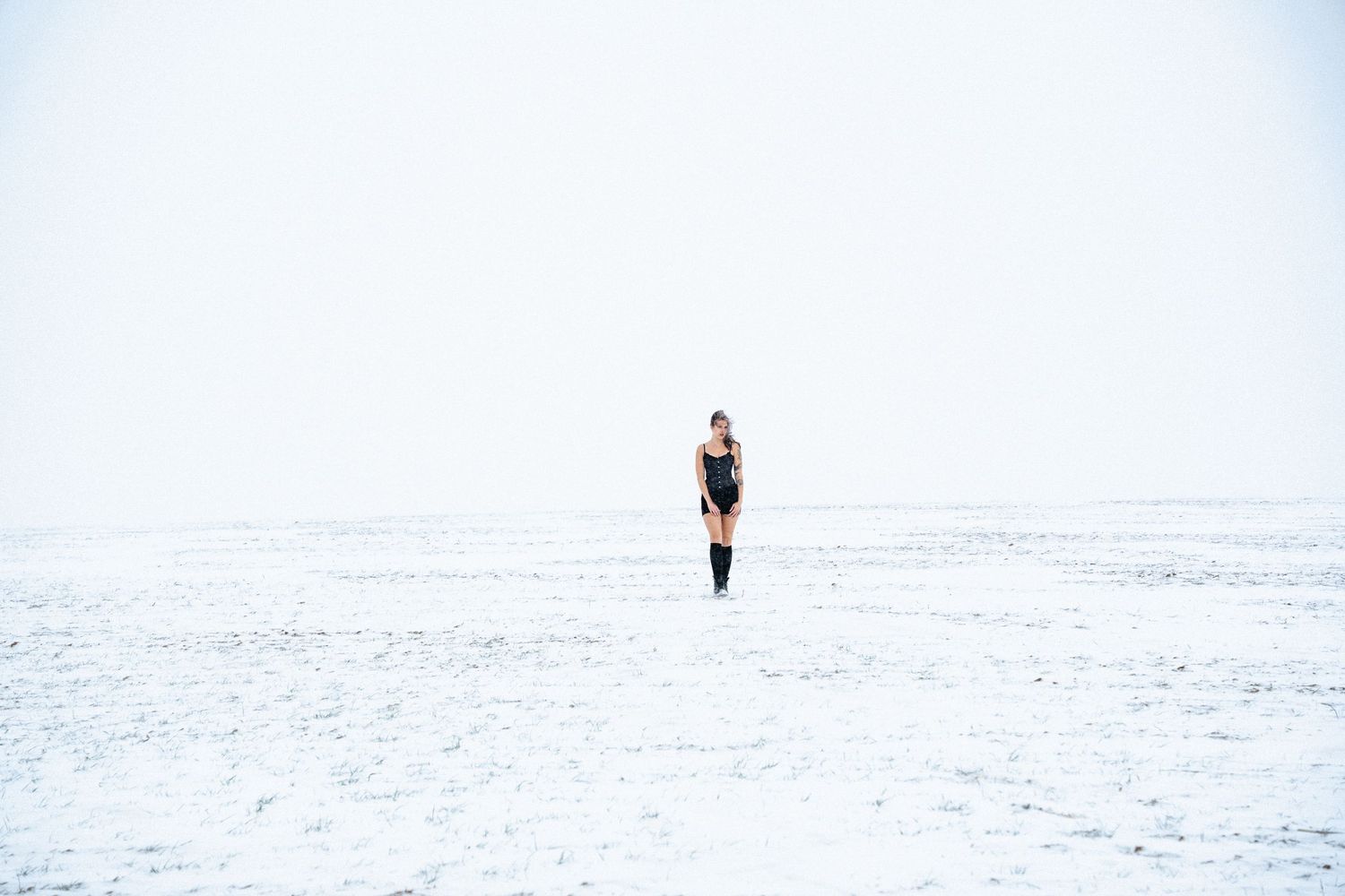 A solitary figure stands in a vast snow-covered landscape against a white winter sky.